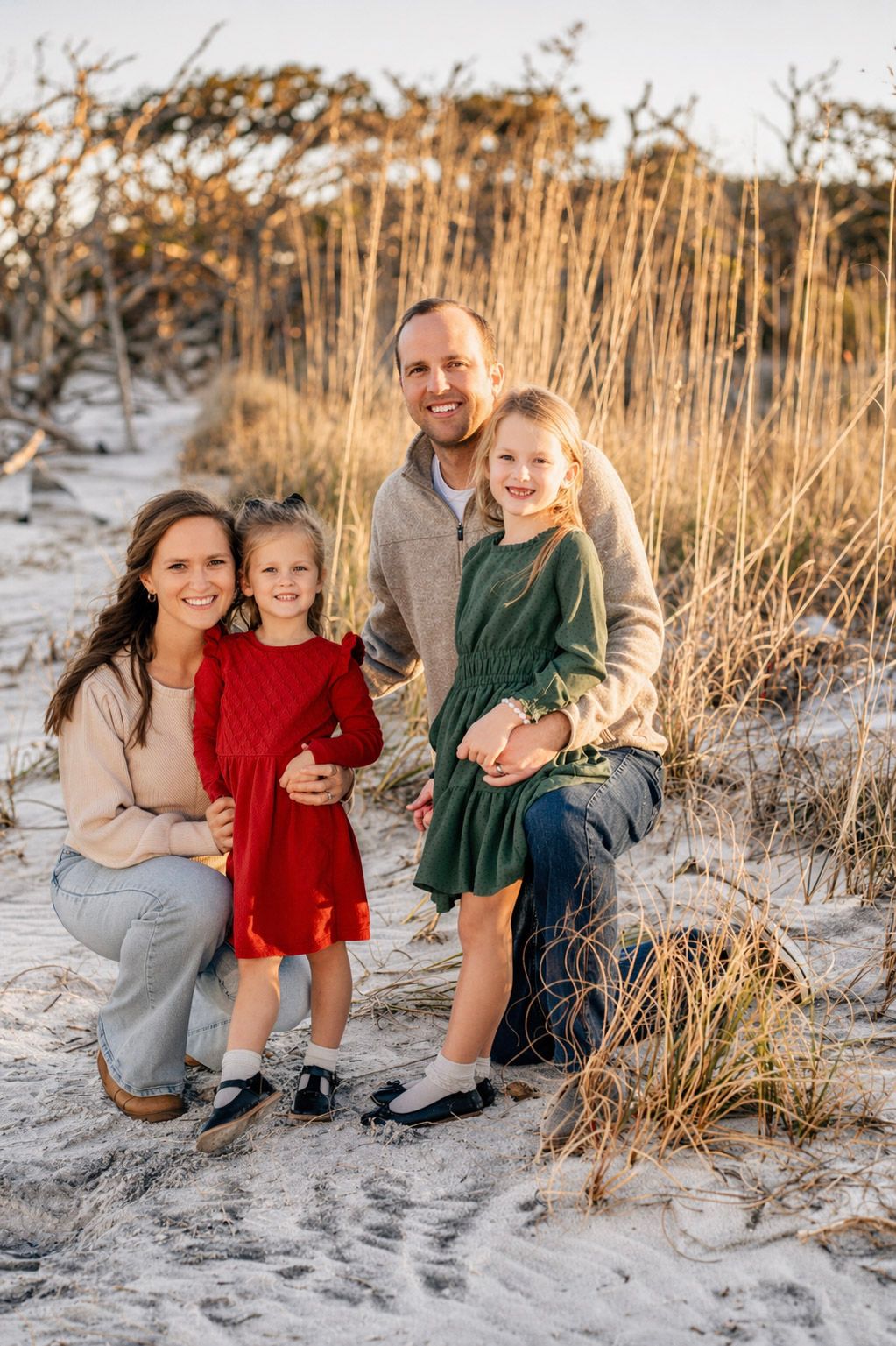 Family of four posing on sandy beach dunes with golden sea oats, two girls in red and green dresses smiling.