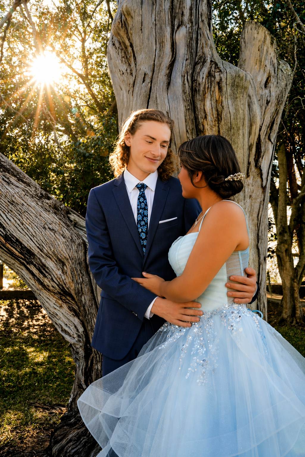 Couple in formal attire embracing near a tree at golden hour, man in navy suit and woman in light blue ball gown.