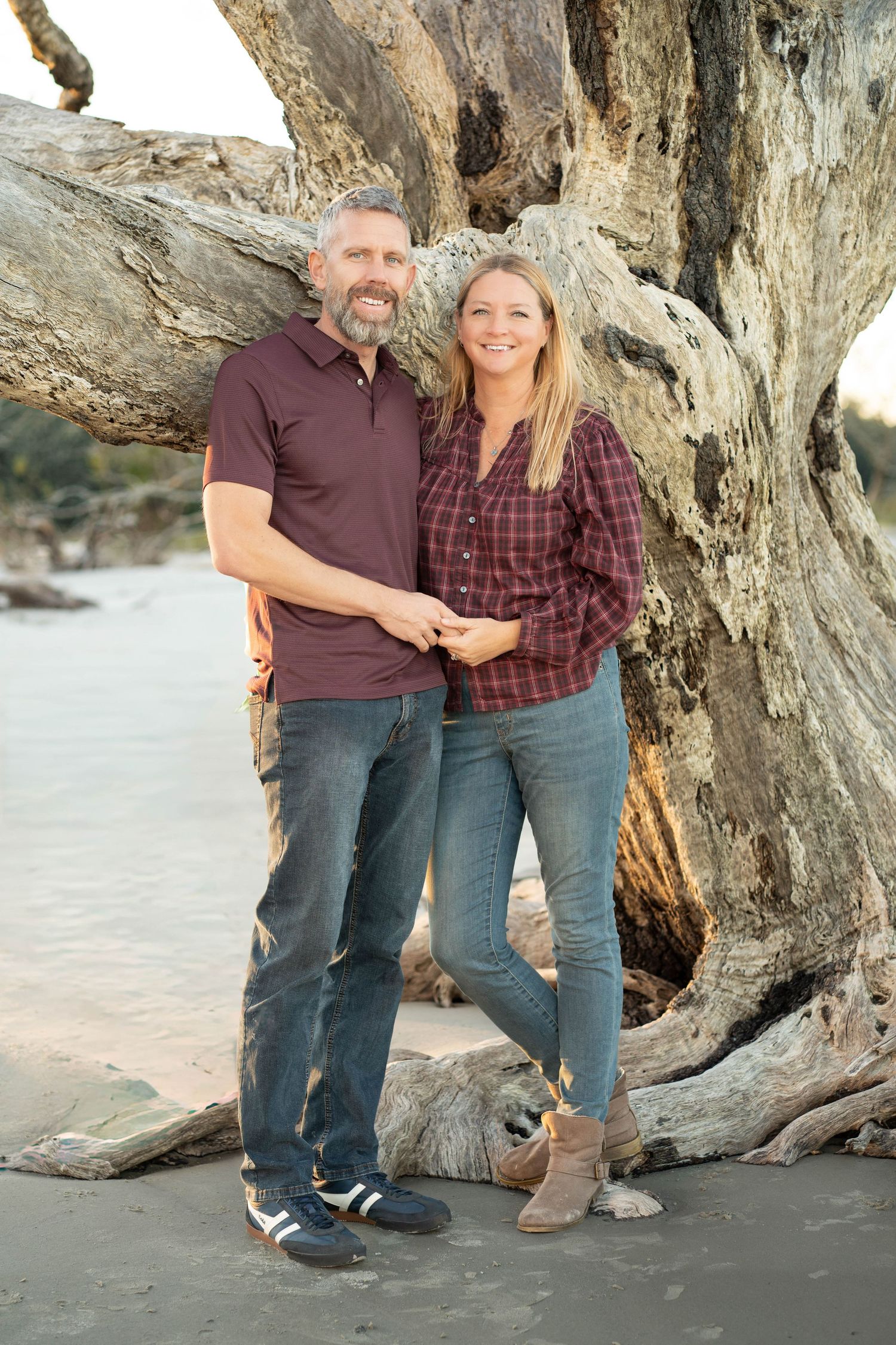 A couple poses together near driftwood and rock formations on a sandy beach at sunset.
