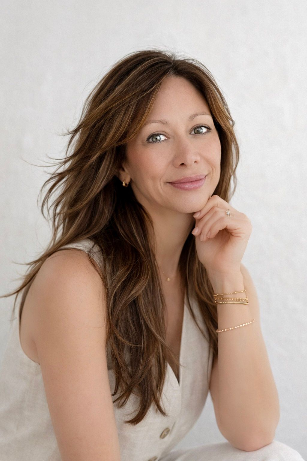 Professional portrait of someone in a white top with long brown hair and delicate gold bracelets against a light background.