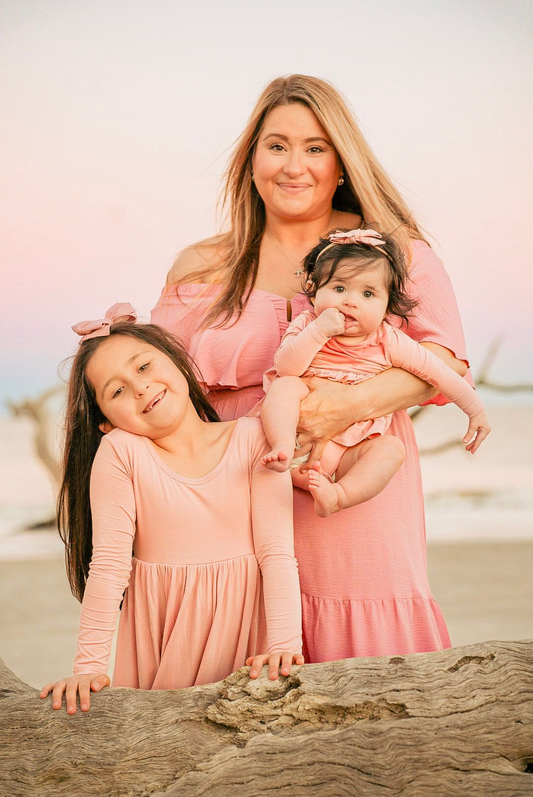 A mother in a pink dress holds her baby while posing with her young daughter on a beach at sunset.