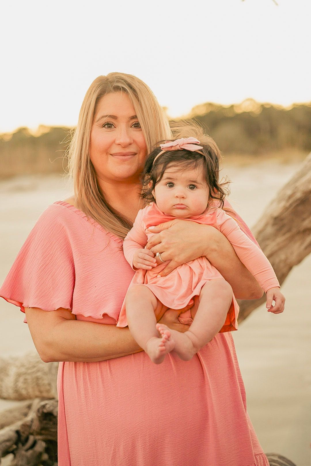 A woman in a coral dress holds her baby girl wearing a matching pink outfit on a sunny beach.