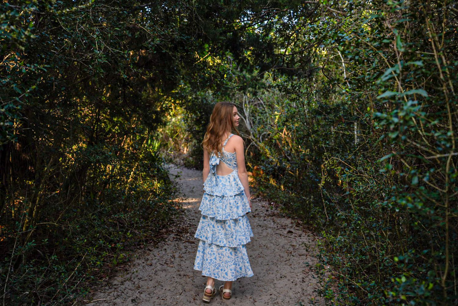 A young girl in a blue ruffled dress walks down a sunlit forest path surrounded by lush green trees.