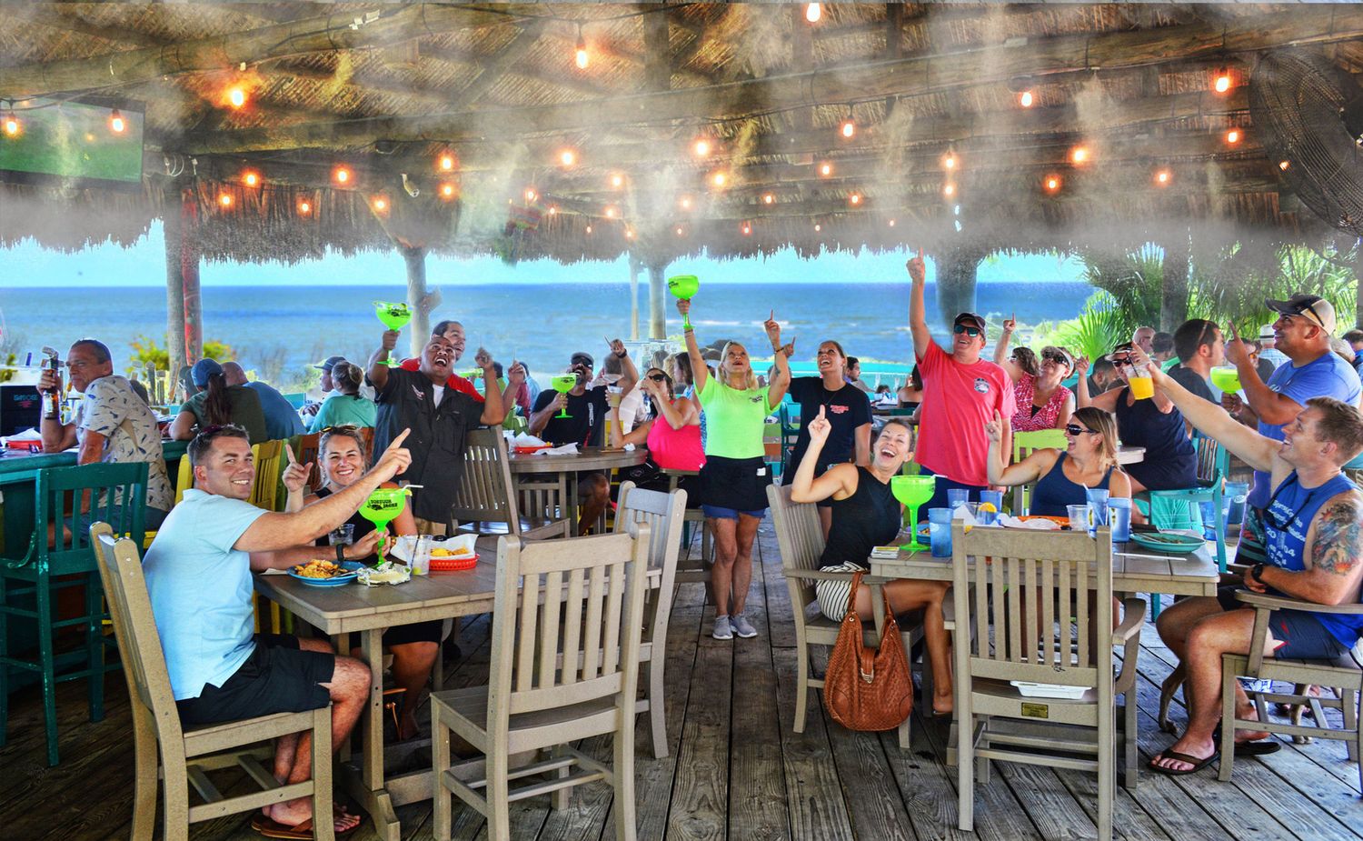 A lively outdoor restaurant deck with string lights and ocean views filled with dining patrons.