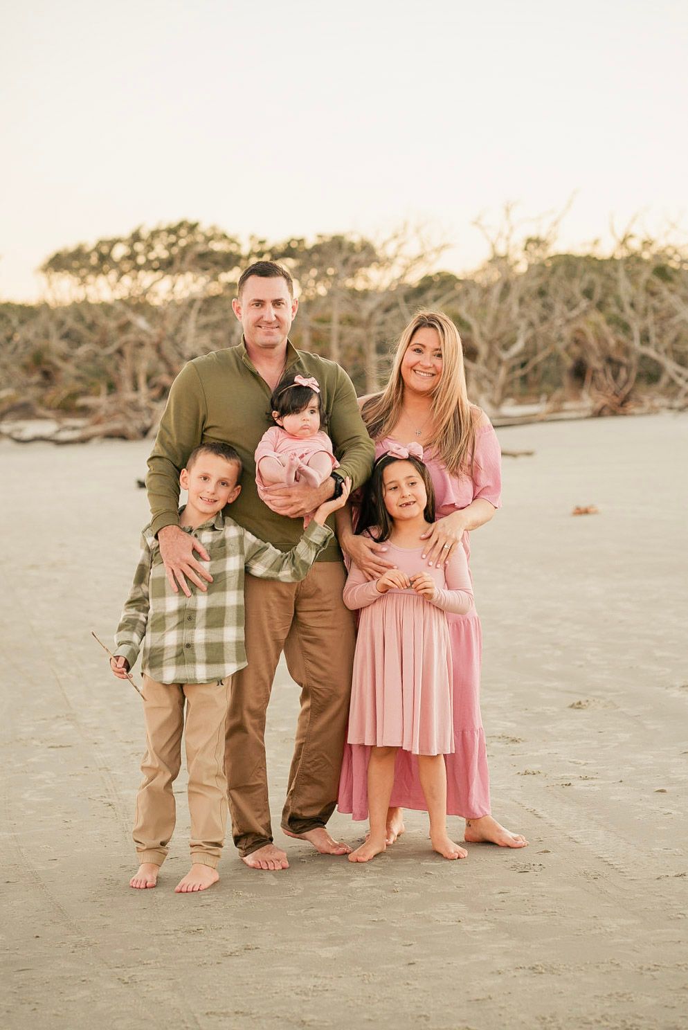 A family poses together barefoot on a beach at sunset wearing coordinated earth-toned and pink outfits.