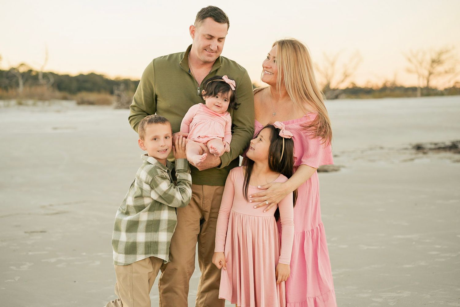 Family of five posing together on a beach at sunset, parents with three children in pink and green outfits.