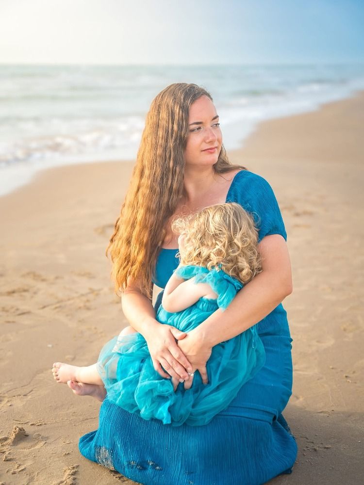 Mother and child in matching turquoise dresses share joyful moments on sunny beach.