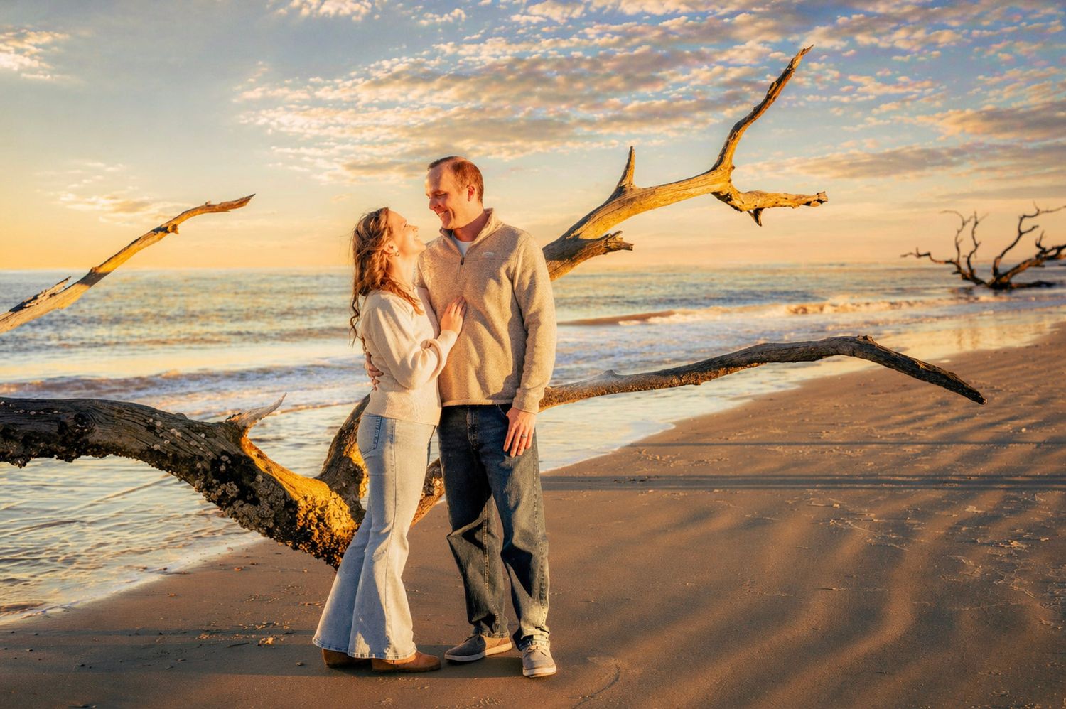 Couple sharing a romantic moment on a sandy beach at sunset with driftwood and colorful sky backdrop.