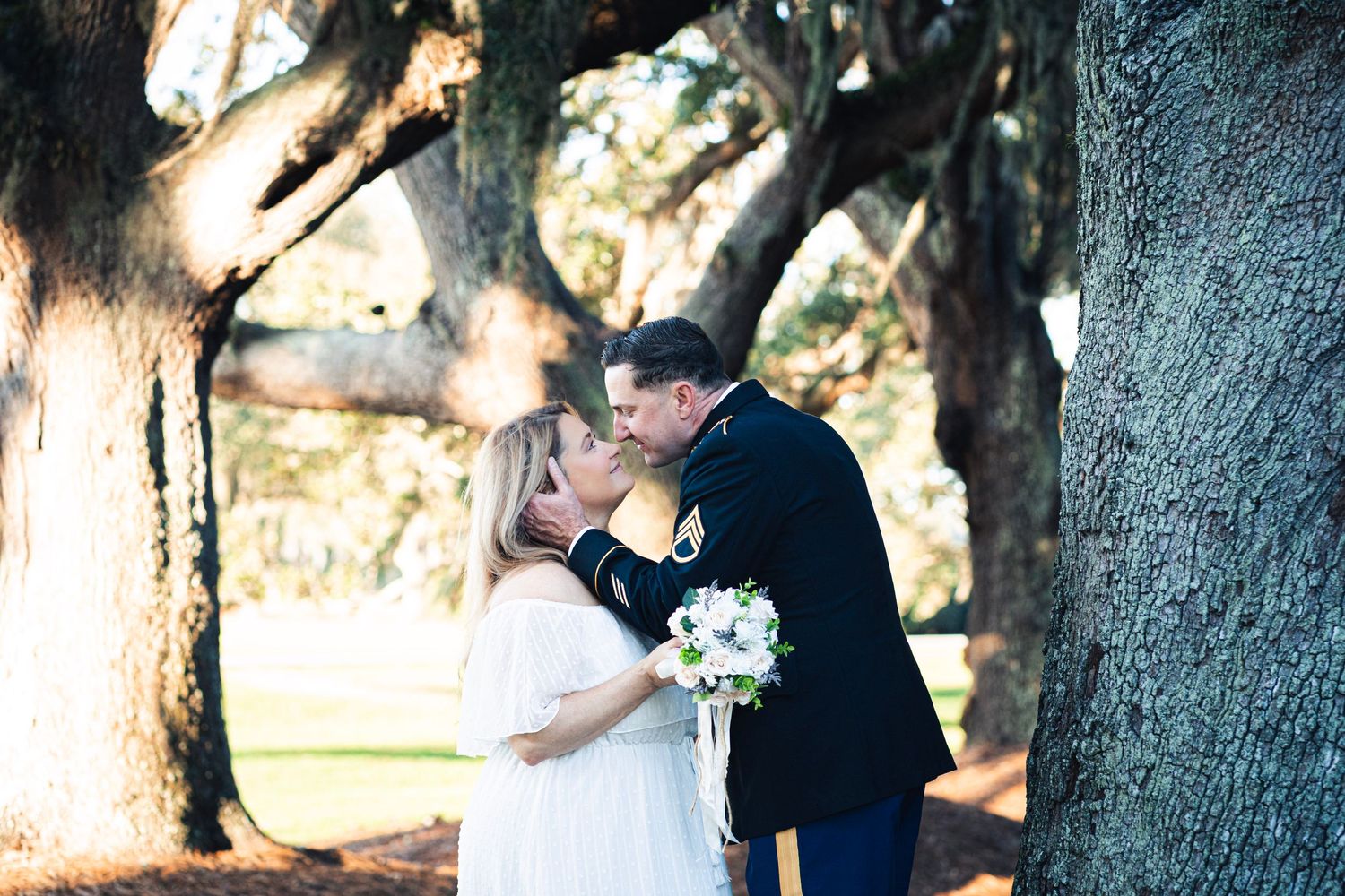 Military groom in dress blues kisses bride in white gown among massive oak trees in romantic outdoor wedding ceremony.
