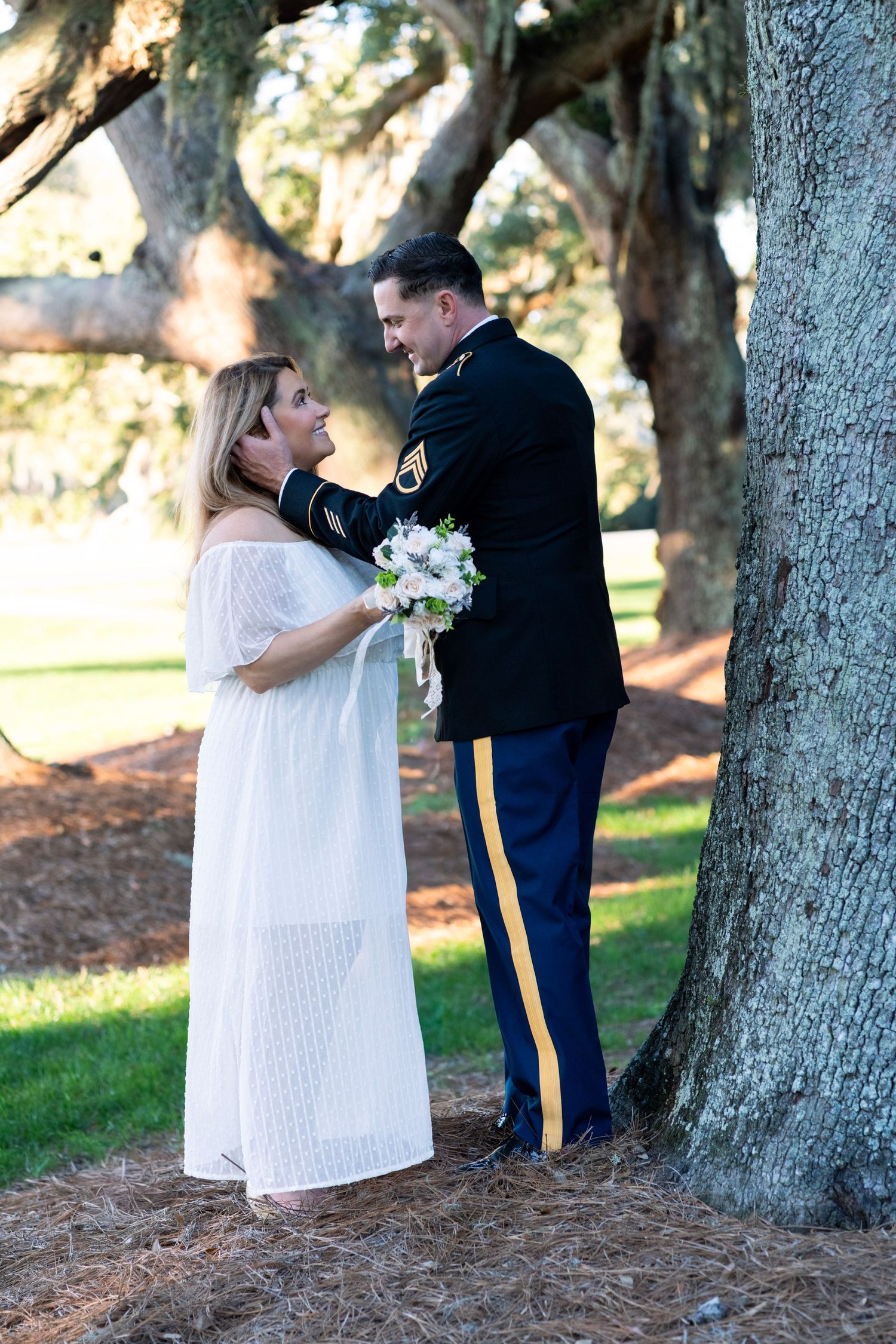 Military groom in dress uniform holds bride in white dress near oak tree during outdoor wedding ceremony.