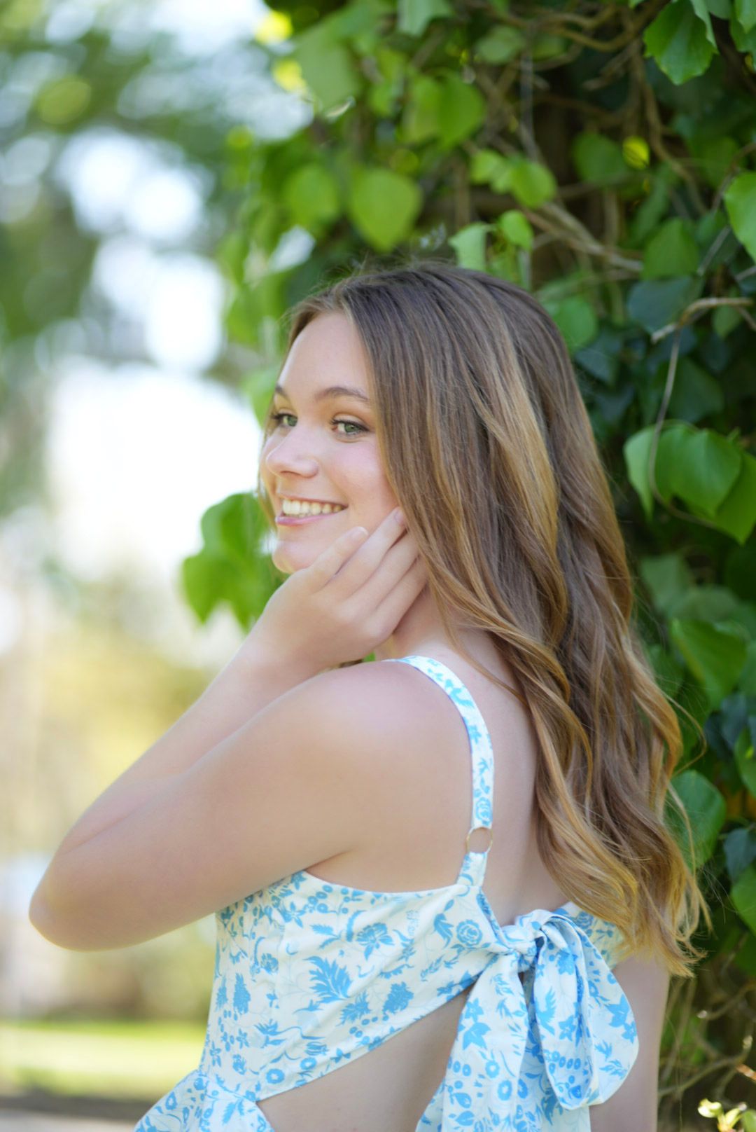Young woman in blue floral sundress smiling over shoulder, long wavy hair, green ivy background.