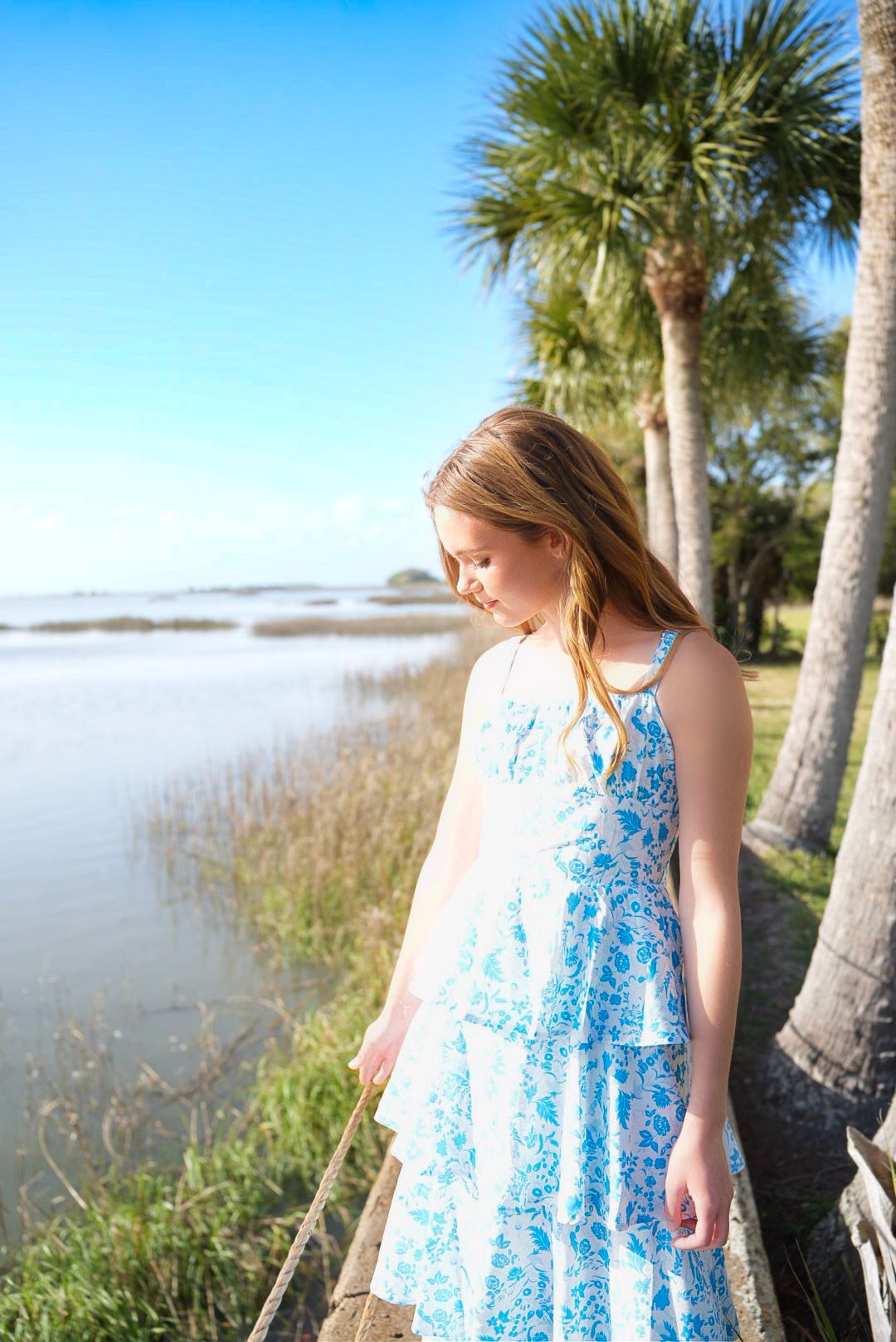 Young girl in blue floral dress standing by a serene waterway with palm trees under a bright blue sky.