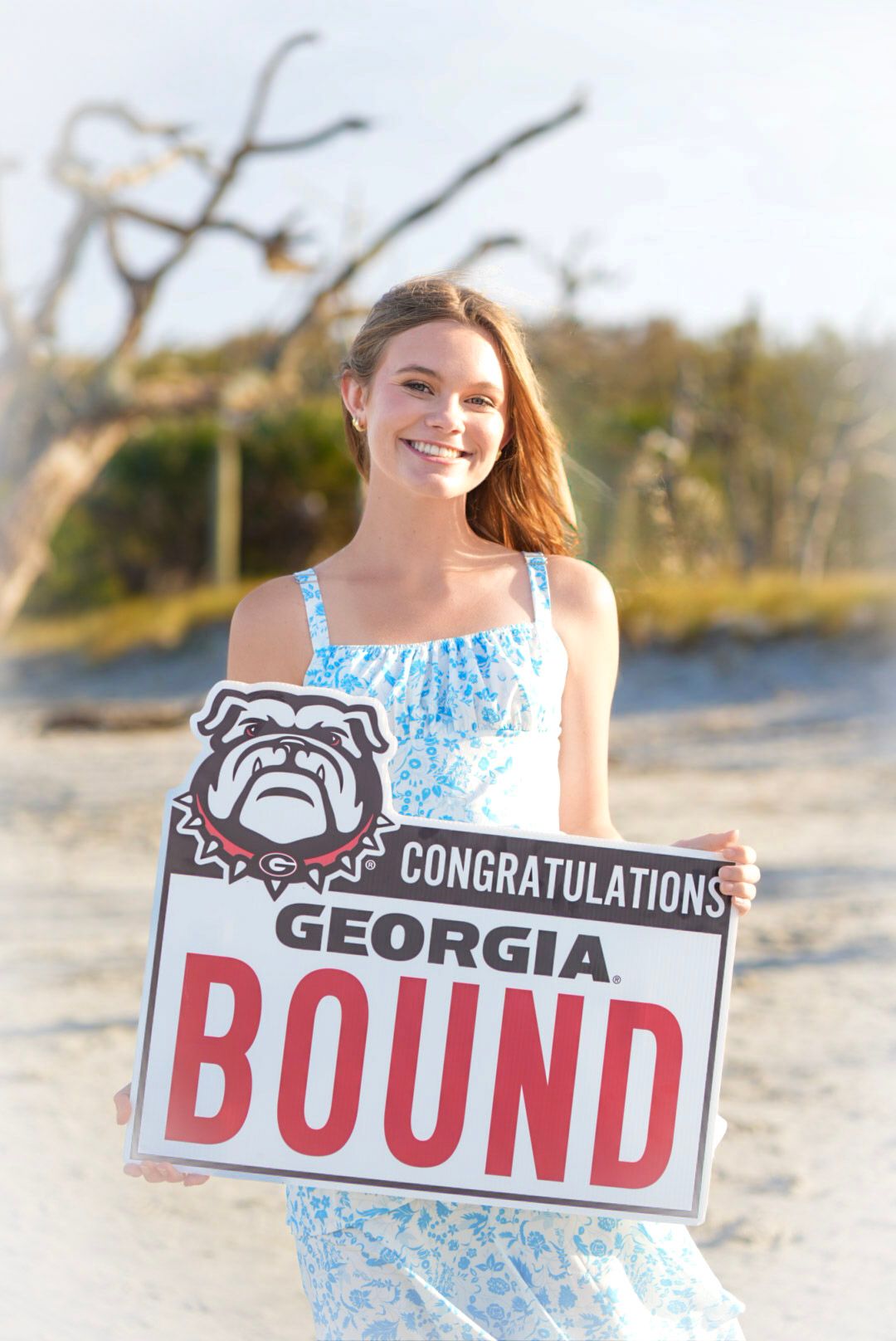 Smiling young woman holding a Congratulations Georgia Bound sign with UGA Bulldogs logo on a sandy beach.