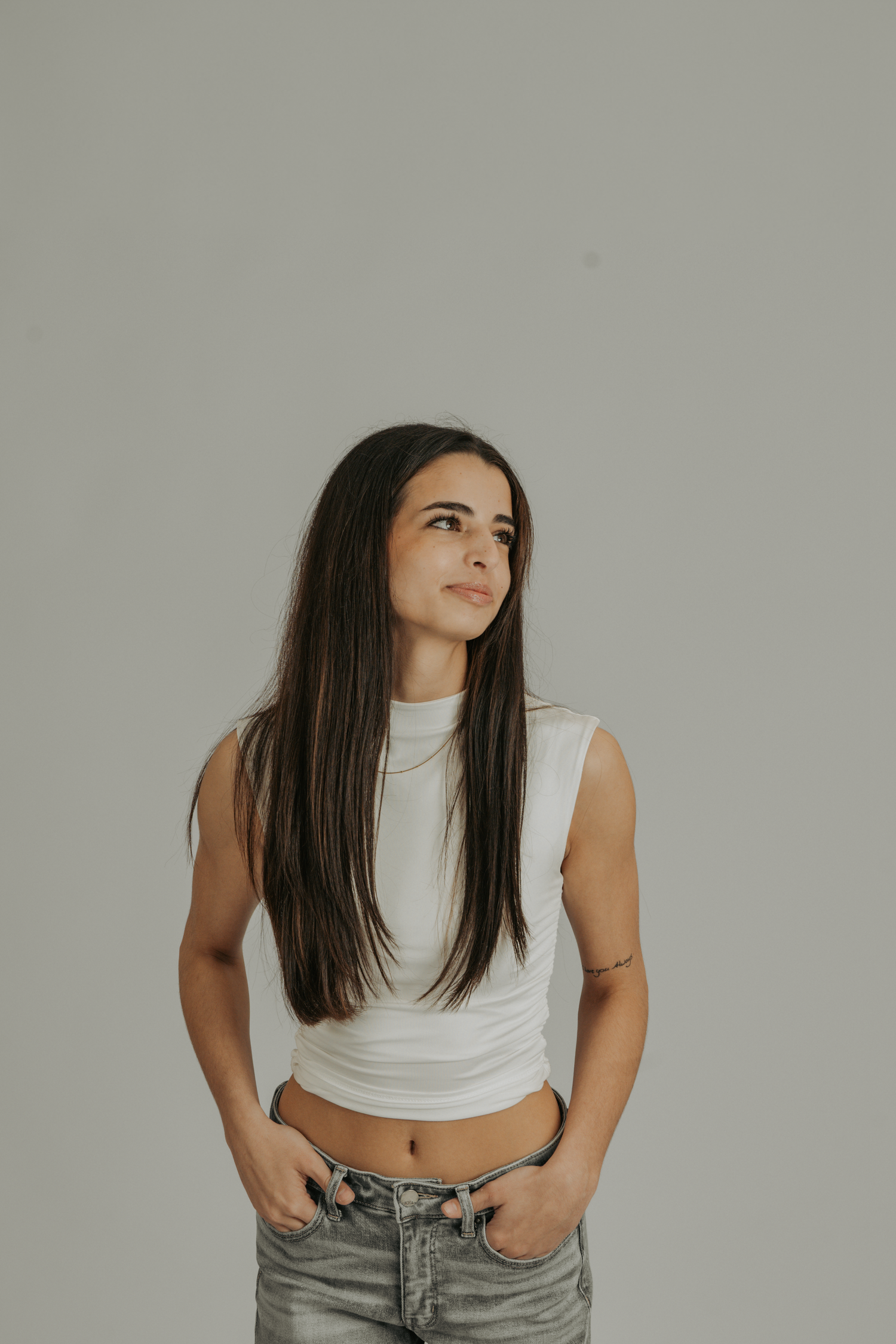 Young woman with long dark hair in white crop top and jeans, hands in pockets, looking up against gray background.
