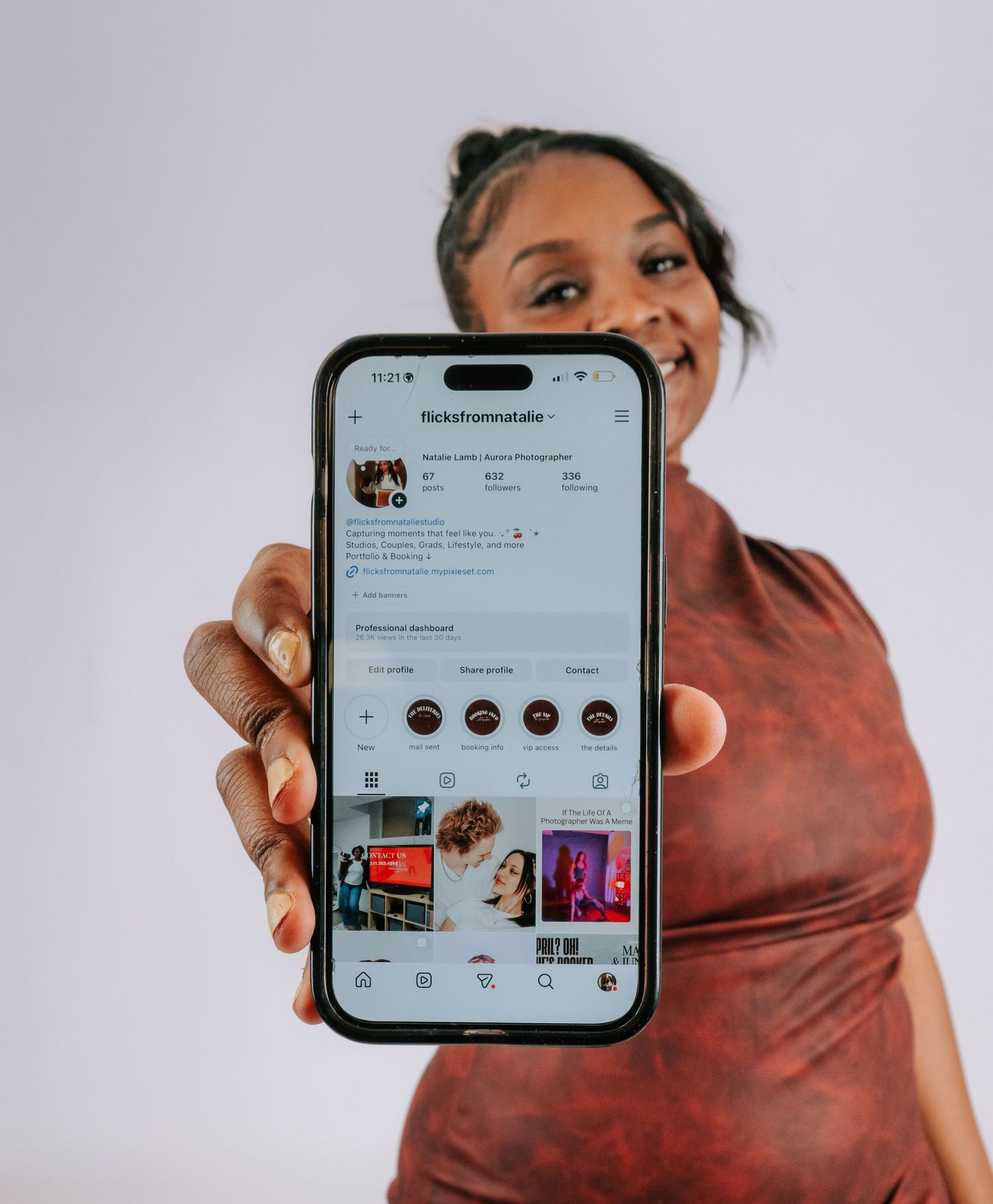 Woman smiling while holding smartphone displaying an Instagram profile page toward camera.