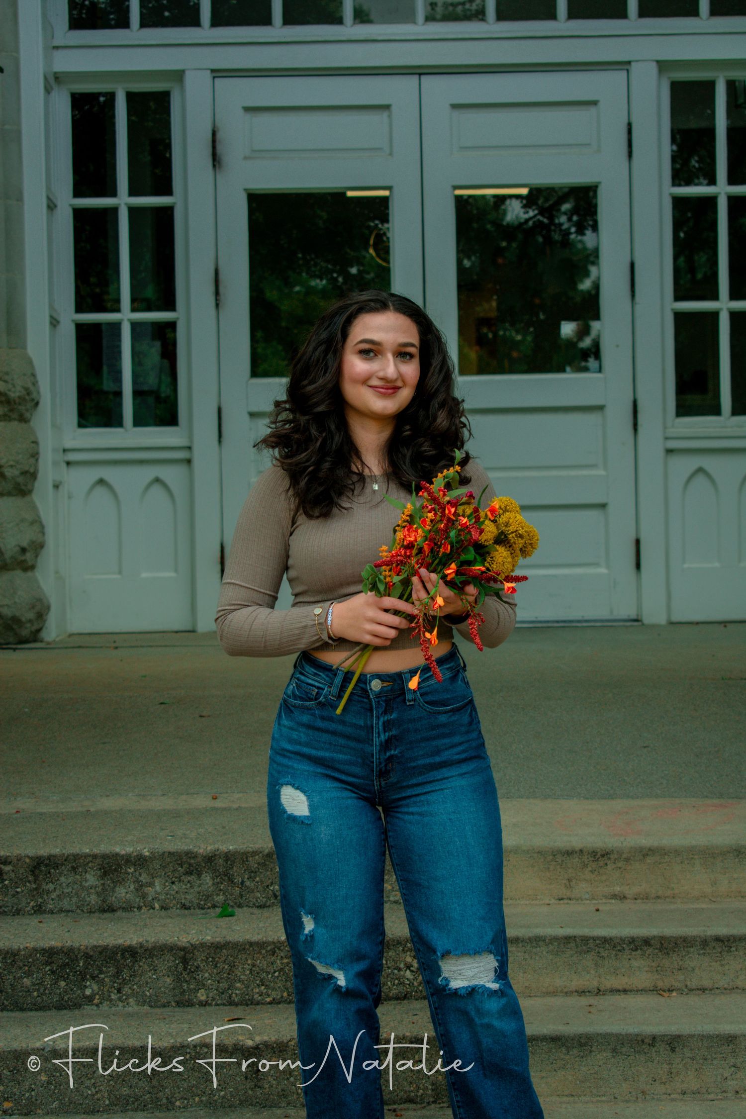 Person in casual outfit holds autumn flowers outside building with white doors.