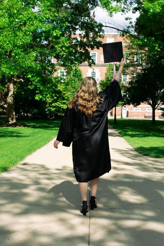 Graduate in black gown walks away on campus path under green trees.