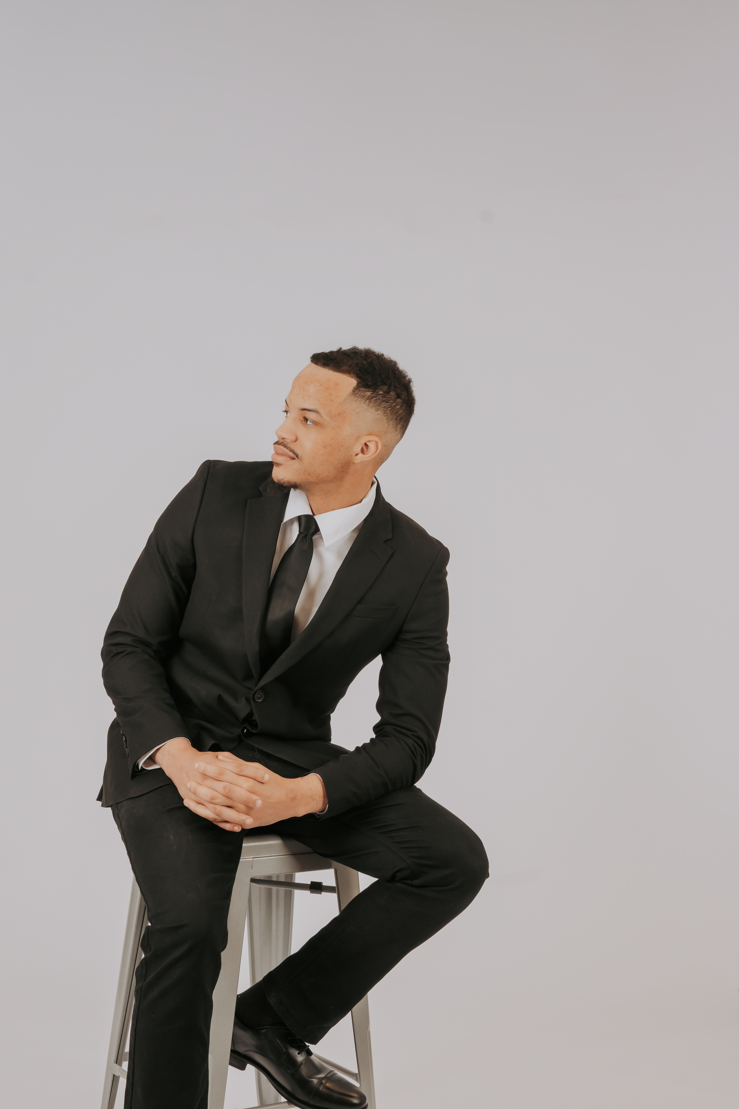 A professional man in a black suit and tie sits on a stool against a gray background, looking confidently to the side.
