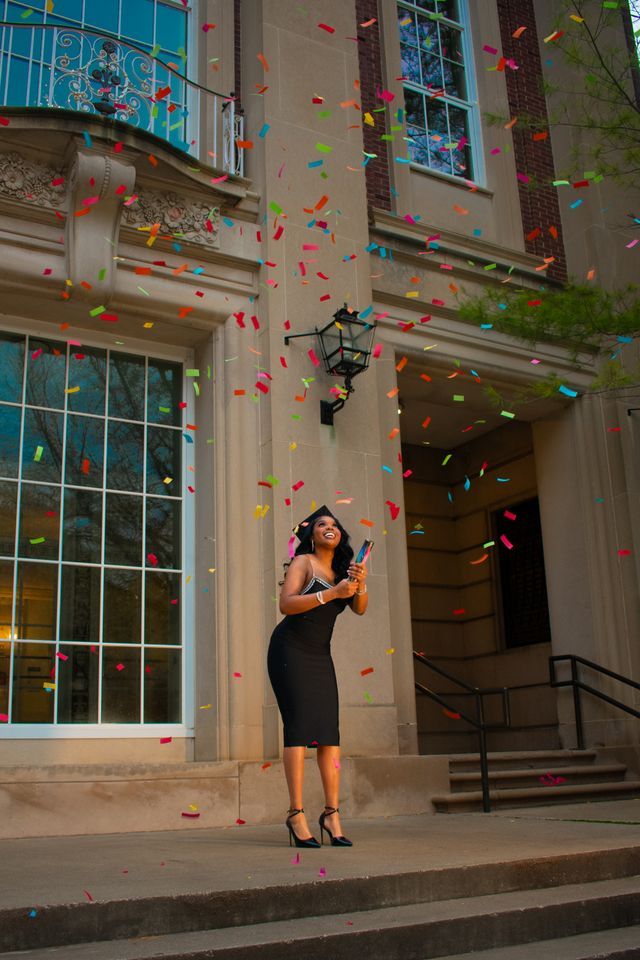 Person in black dress celebrates with colorful confetti outside building entrance.