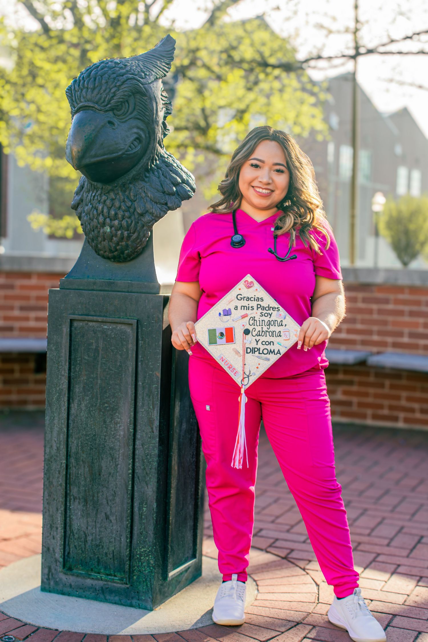 Healthcare professional in bright pink scrubs poses with decorative graduation cap next to campus mascot statue.