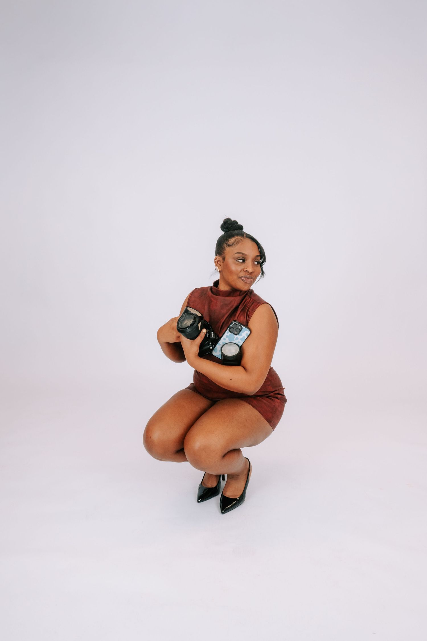 Woman in brown dress crouching in heels, holding vintage cameras, posing against white background.