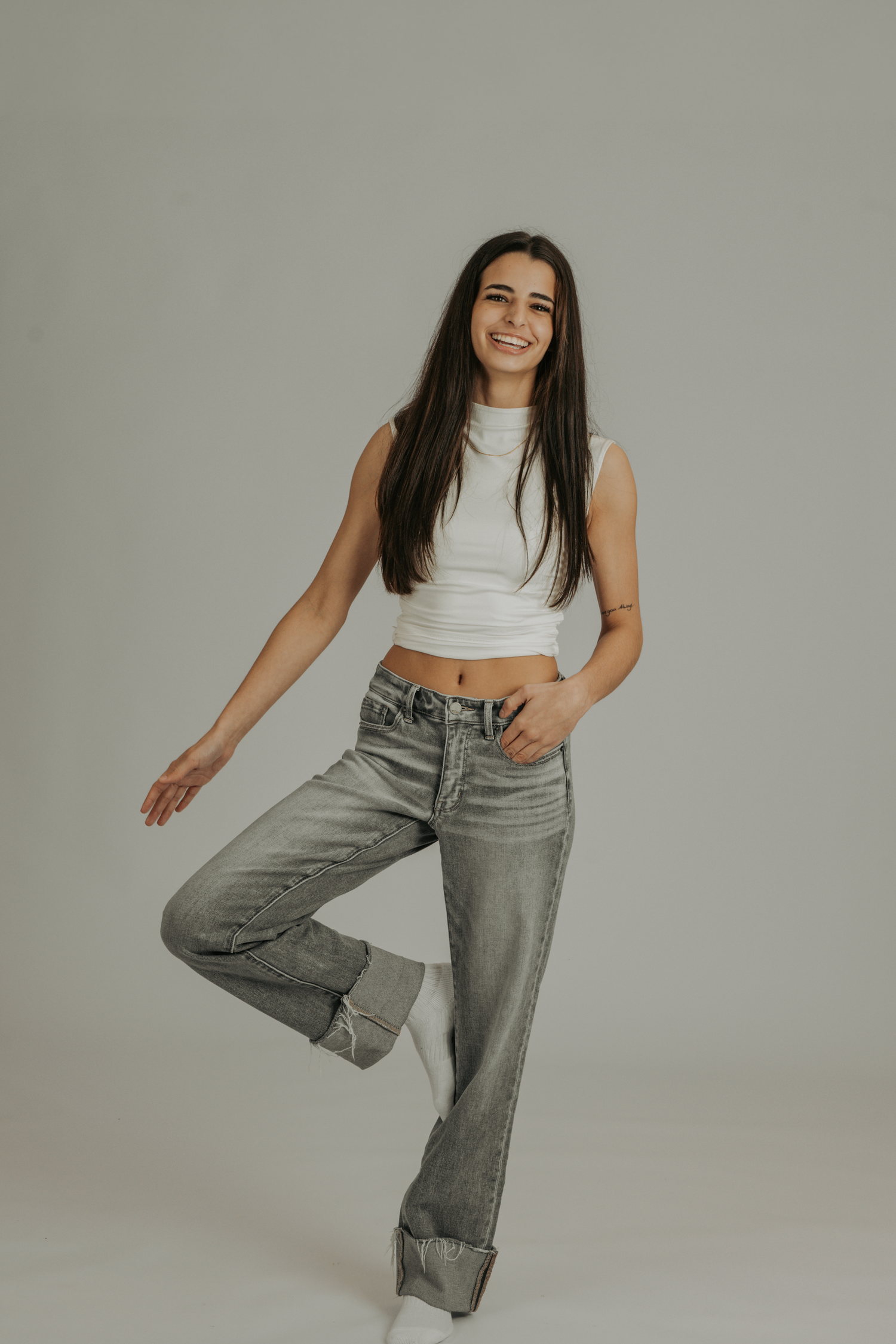 Smiling young woman in white crop top and gray jeans posing on one leg against a light gray background.