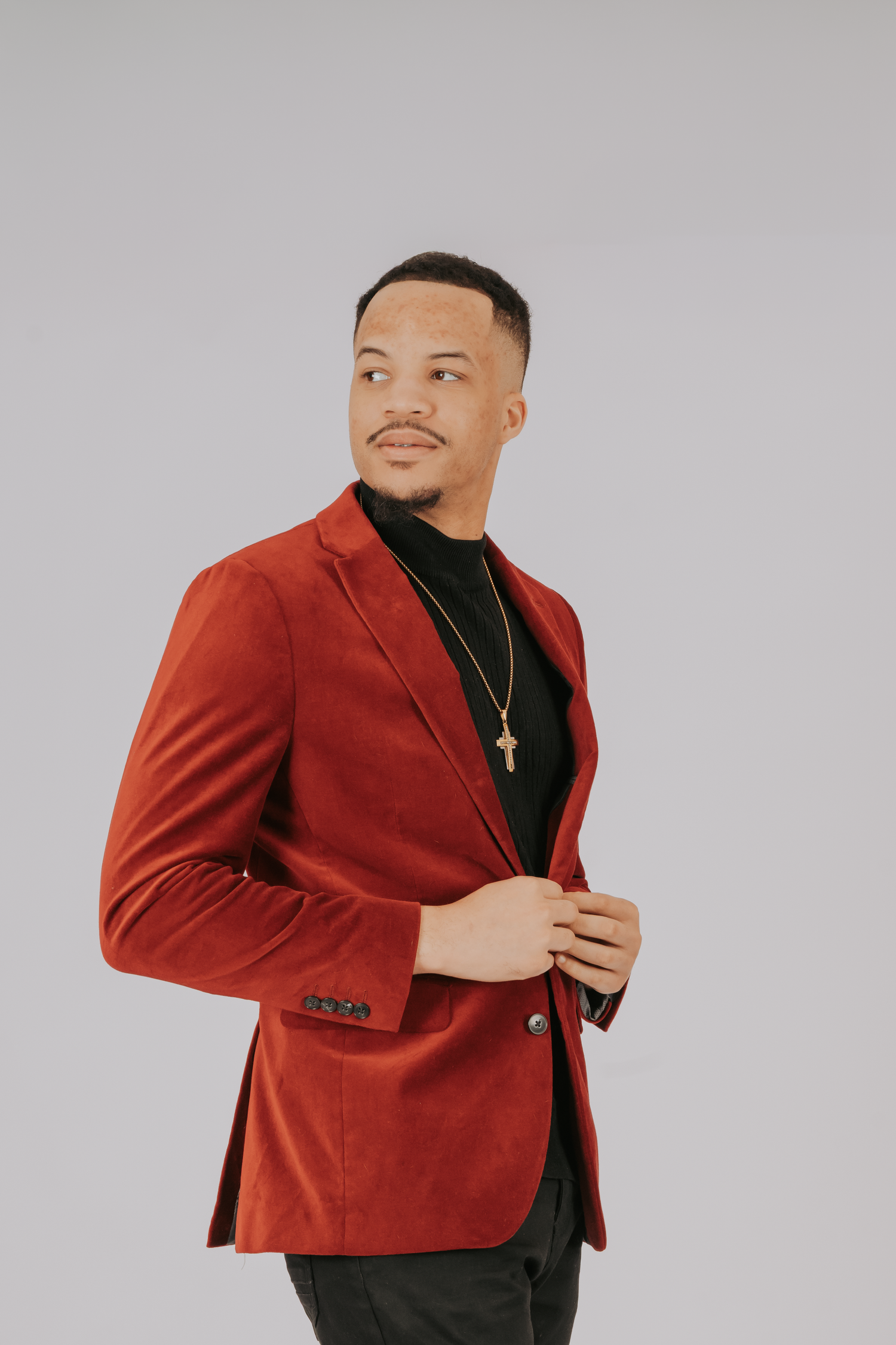 Man in red velvet blazer and black turtleneck wearing gold cross necklace posing confidently against gray background.