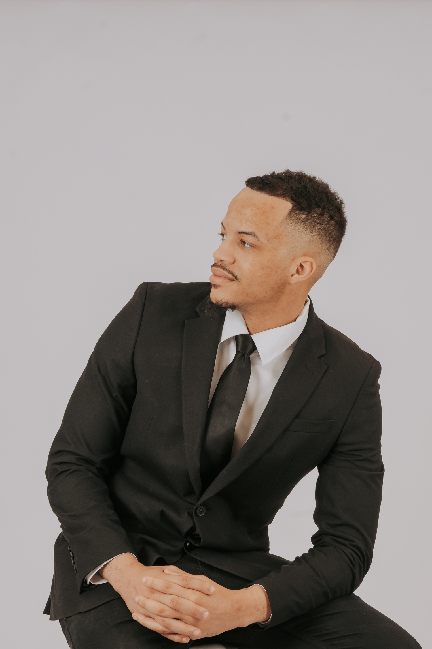 A young man in a black suit and tie sits confidently with hands clasped, gazing to the side against a gray background.