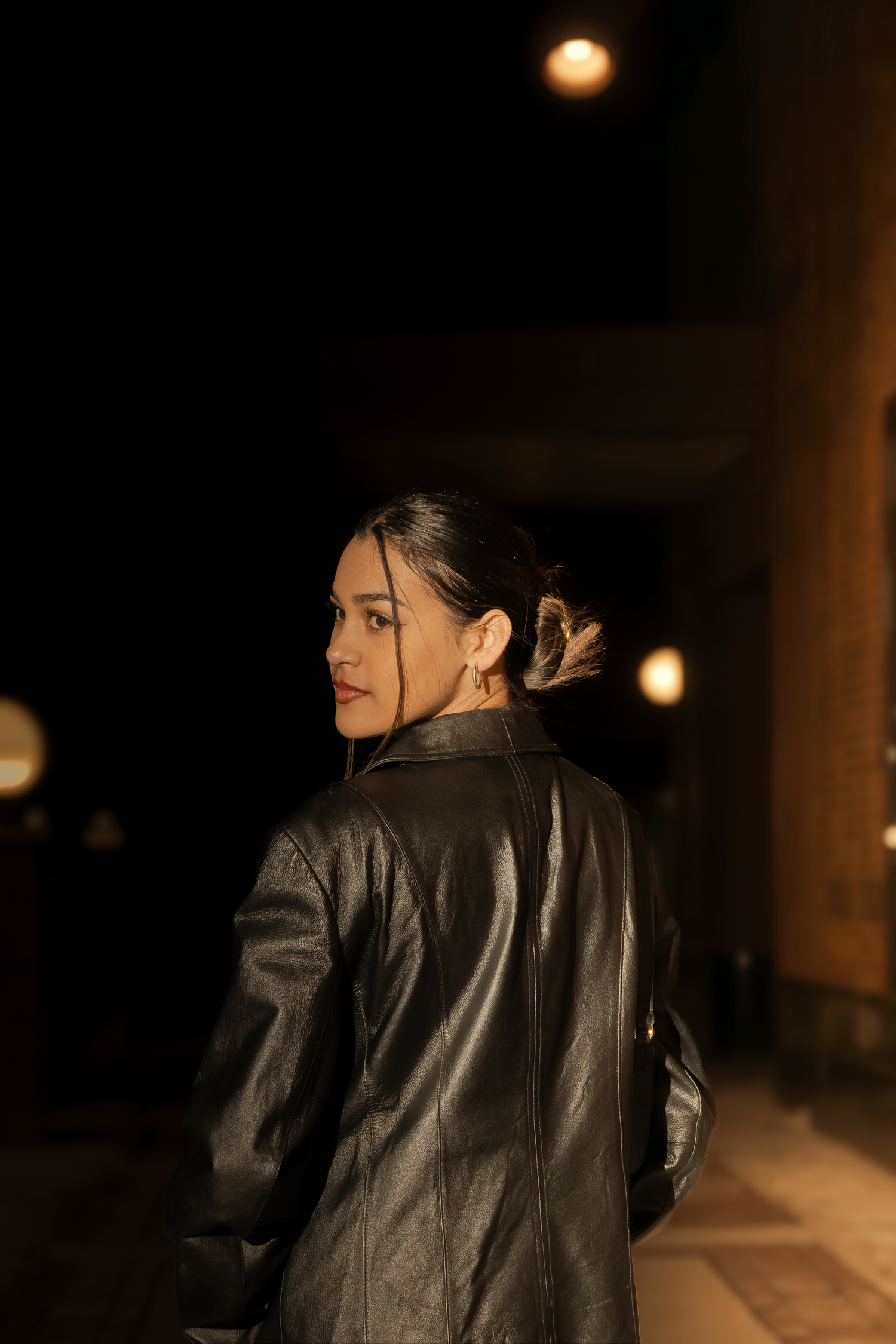 Woman in leather jacket walking away in dimly lit hallway with ambient lighting.