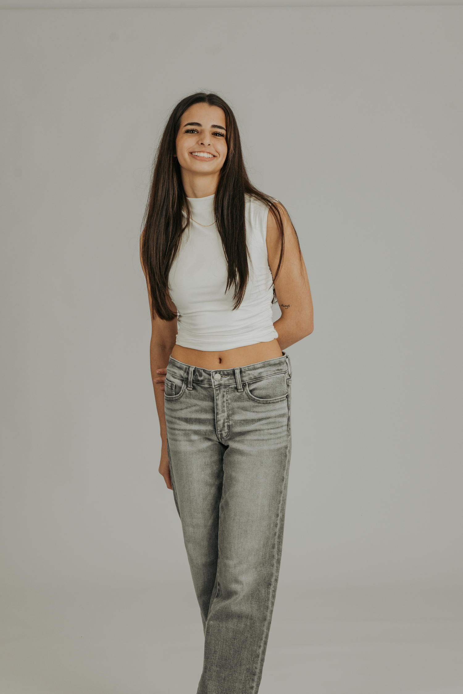 Young woman smiling in white crop top and gray jeans, posing against a light gray background.