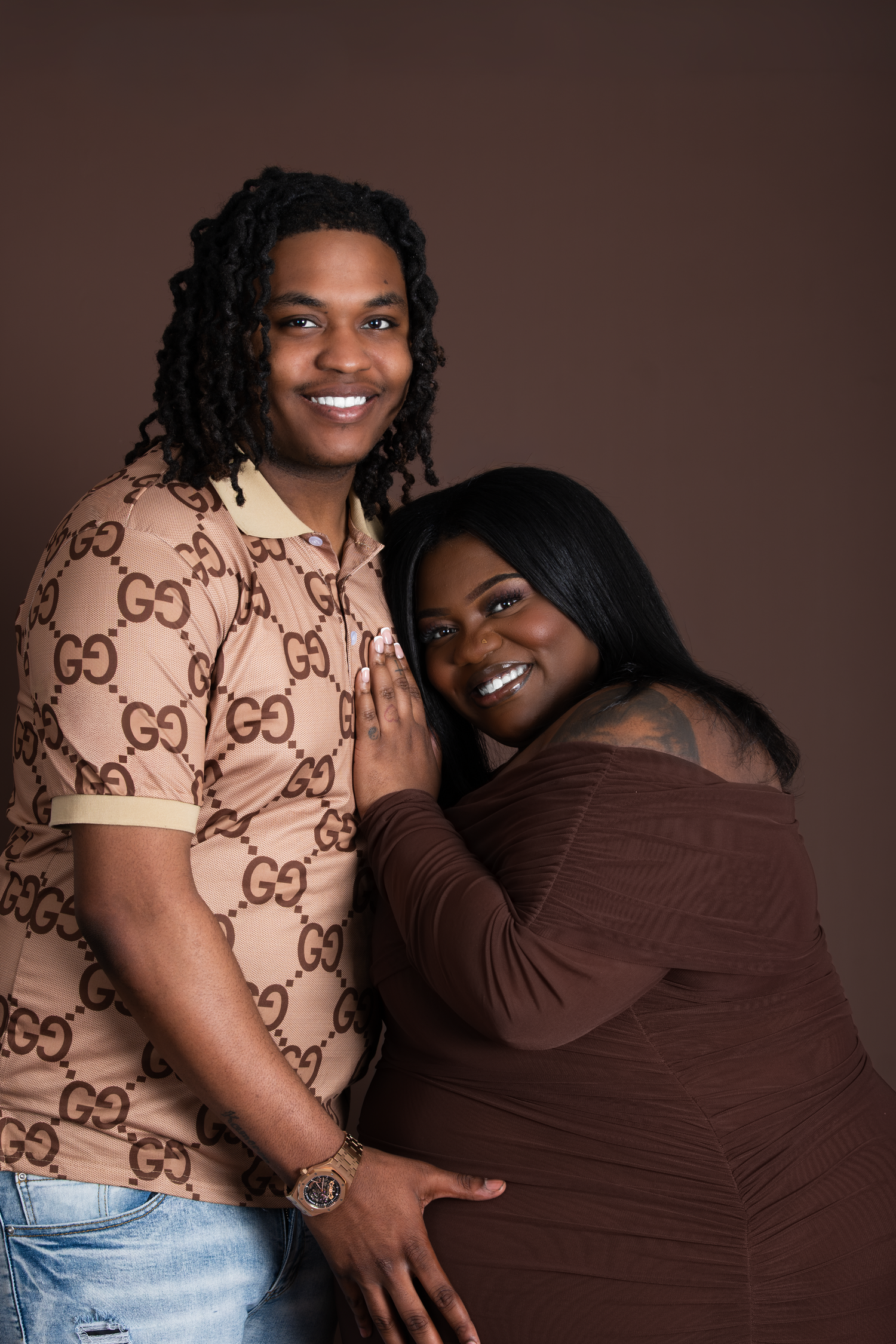 A smiling couple poses together in a studio, man in Gucci shirt and woman in brown dress against brown background.