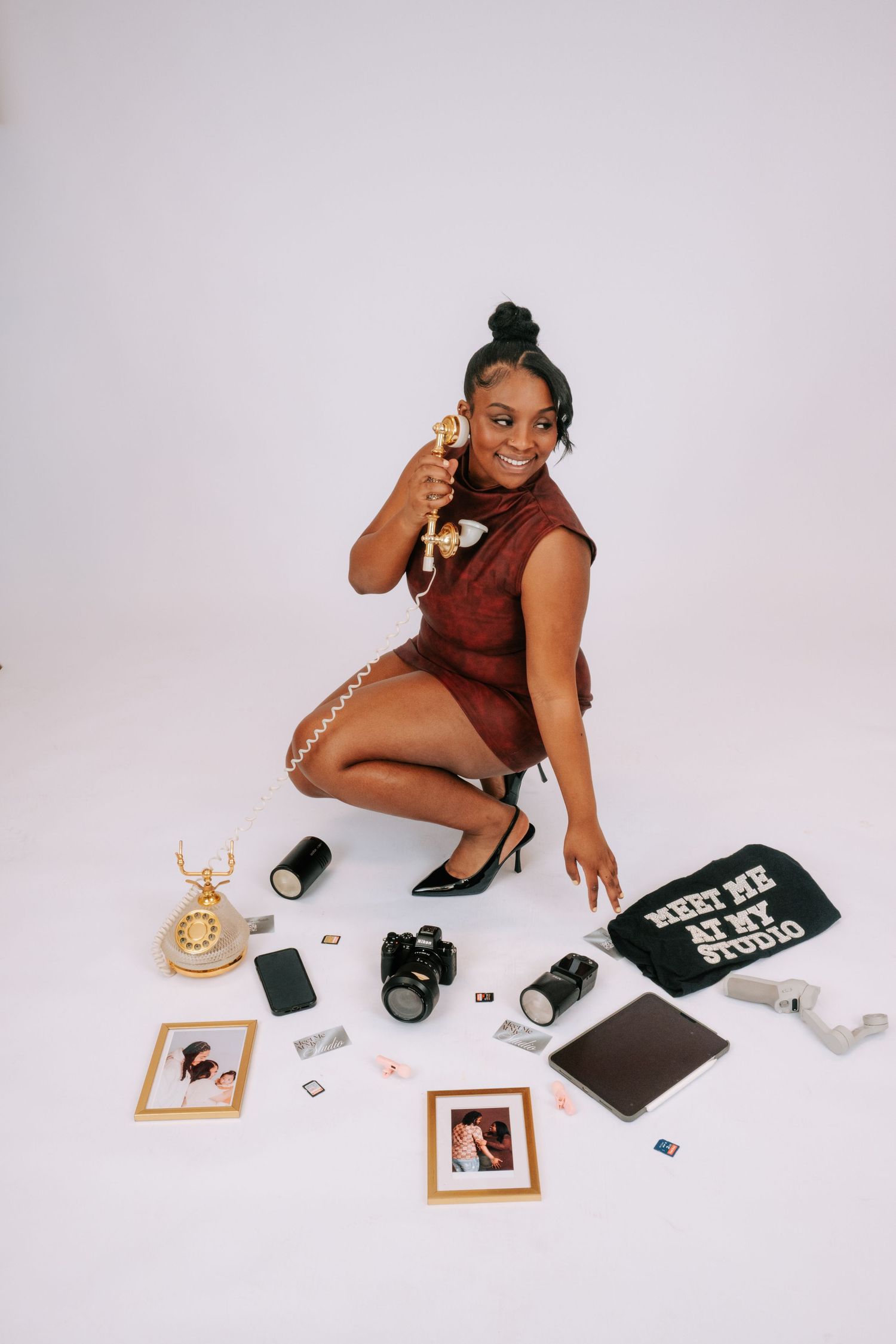Young woman in burgundy dress poses on white studio floor surrounded by camera, photos, and vintage phone props.