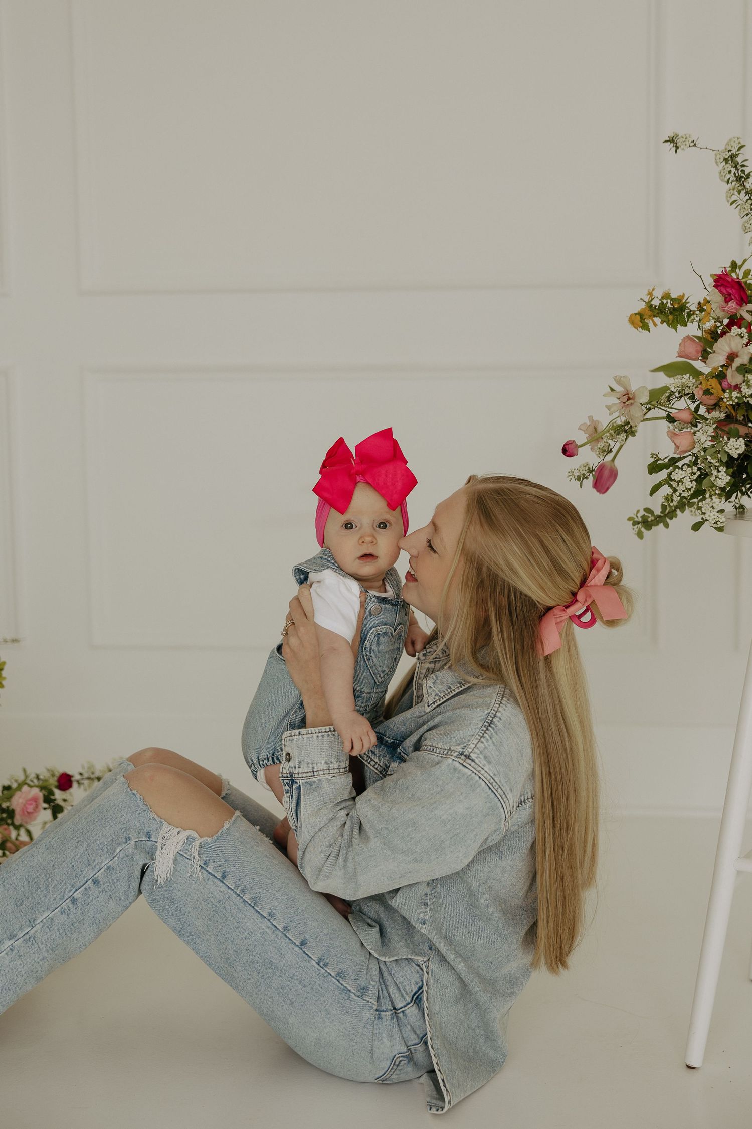 Parent and child in denim share playful moment on floor with pink bow accessories.