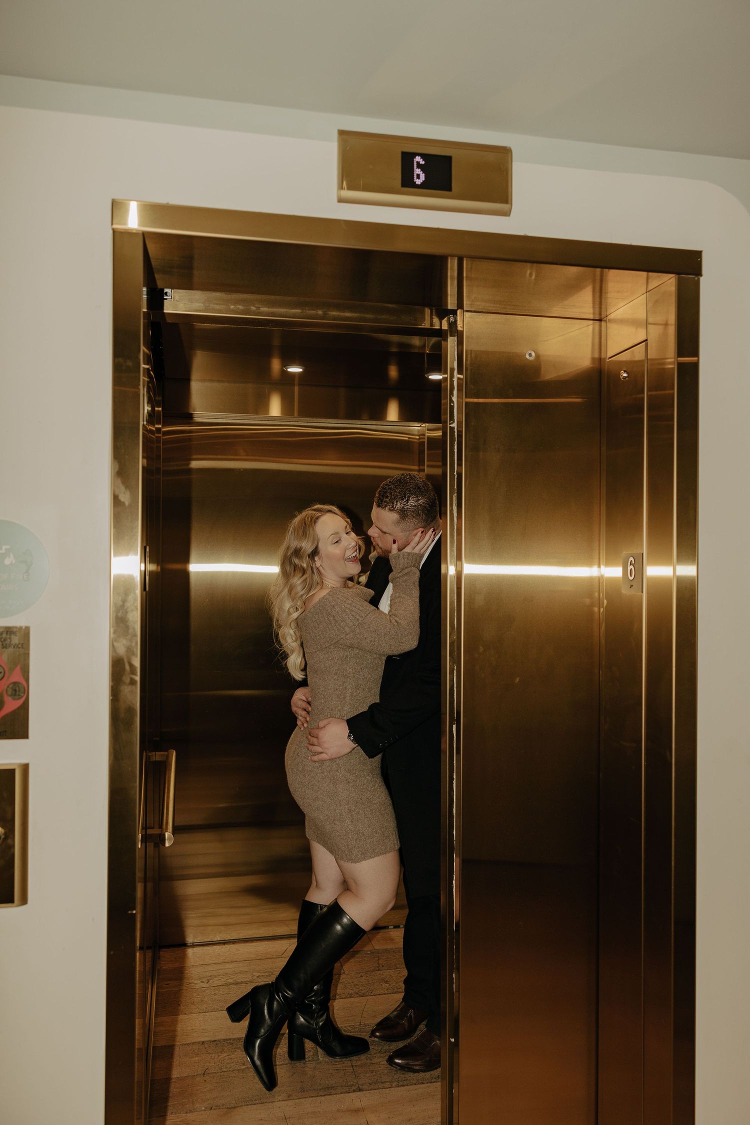 A couple embraces romantically by gold-toned elevator doors in a modern building.