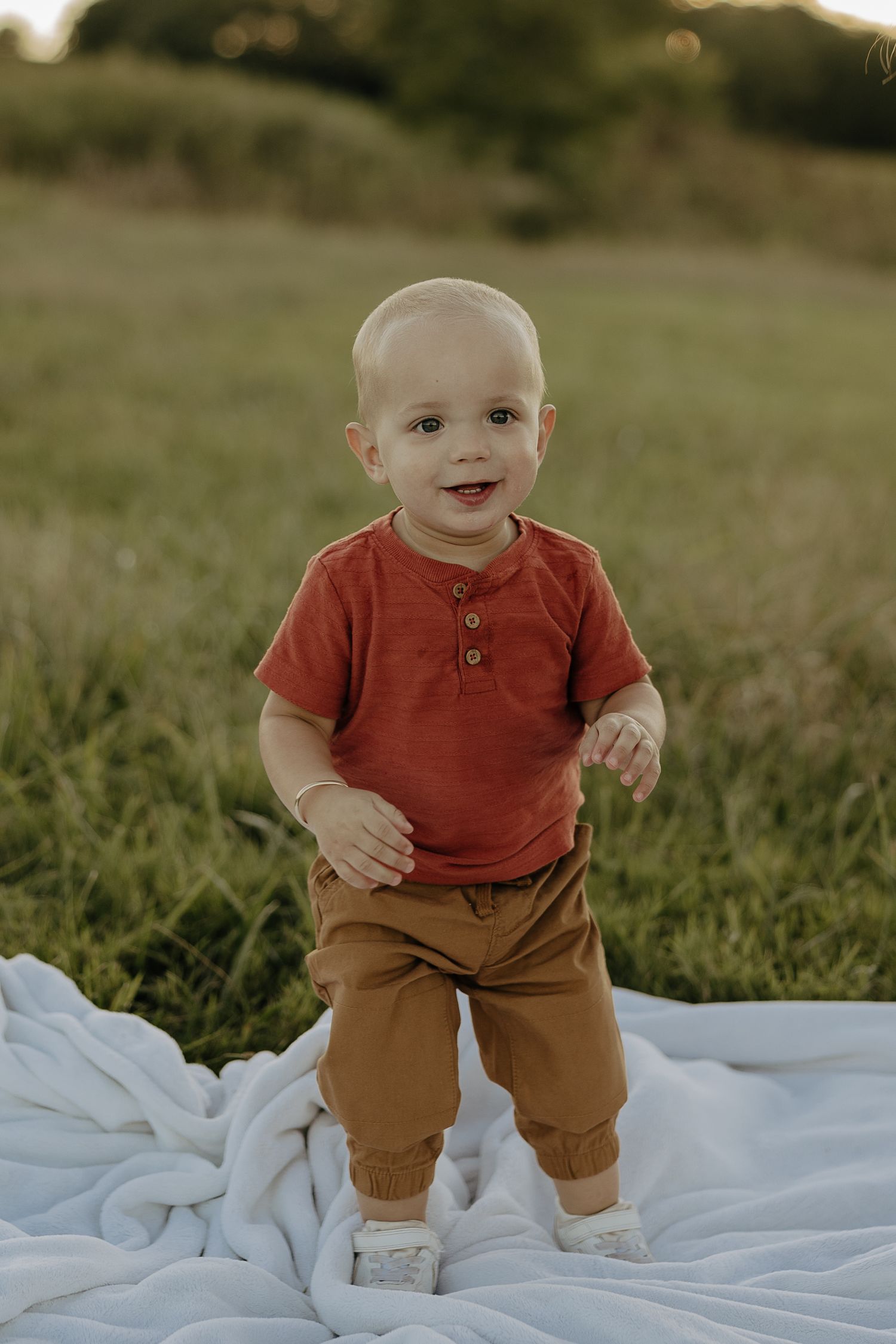 Baby wearing red shirt and brown pants stands on white blanket in grassy field.