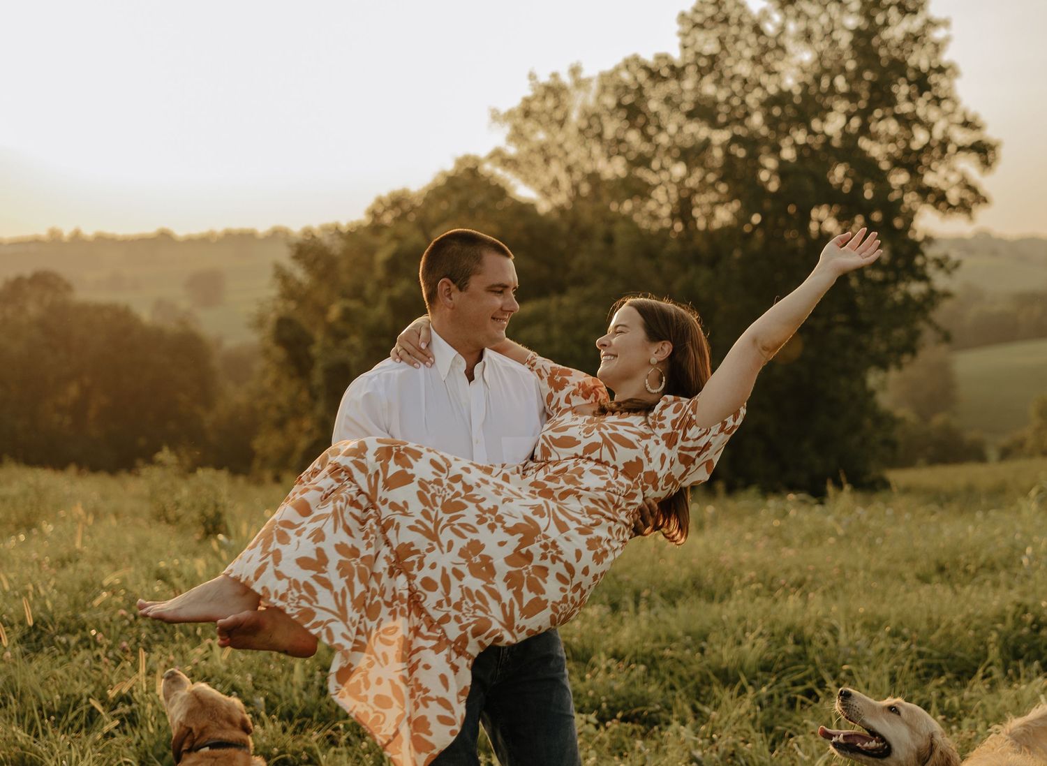 Playful couple dances in golden sunlit field with dog watching nearby.