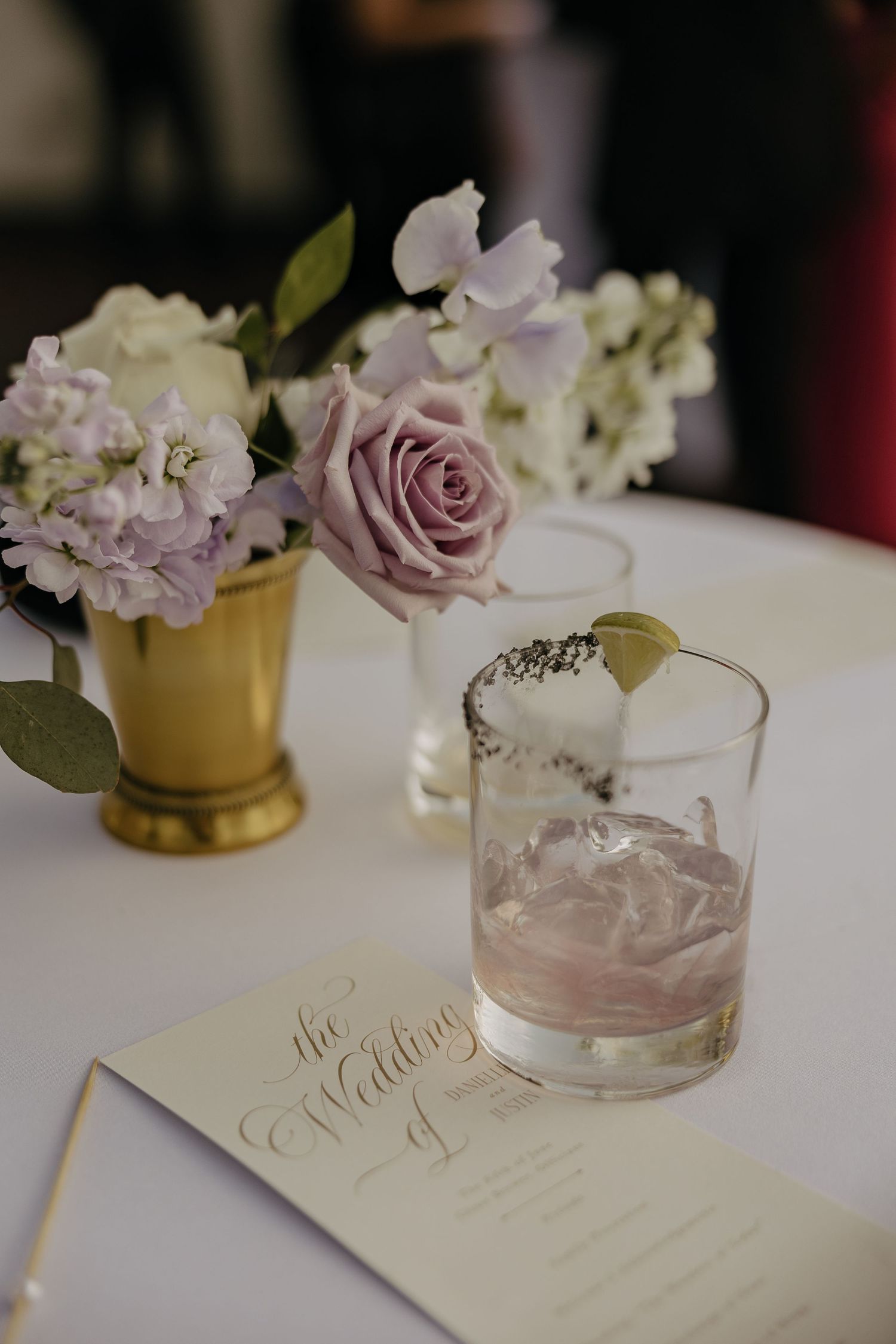 Wedding reception details with pink roses, gold vase, and cocktail glass on white linen.