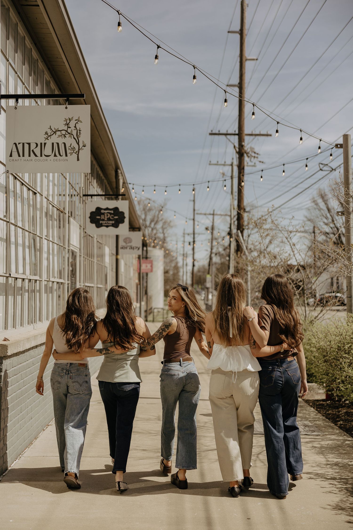 Five women with long hair walking together past the Atrium storefront on a sunny day, wearing jeans and stylish tops.