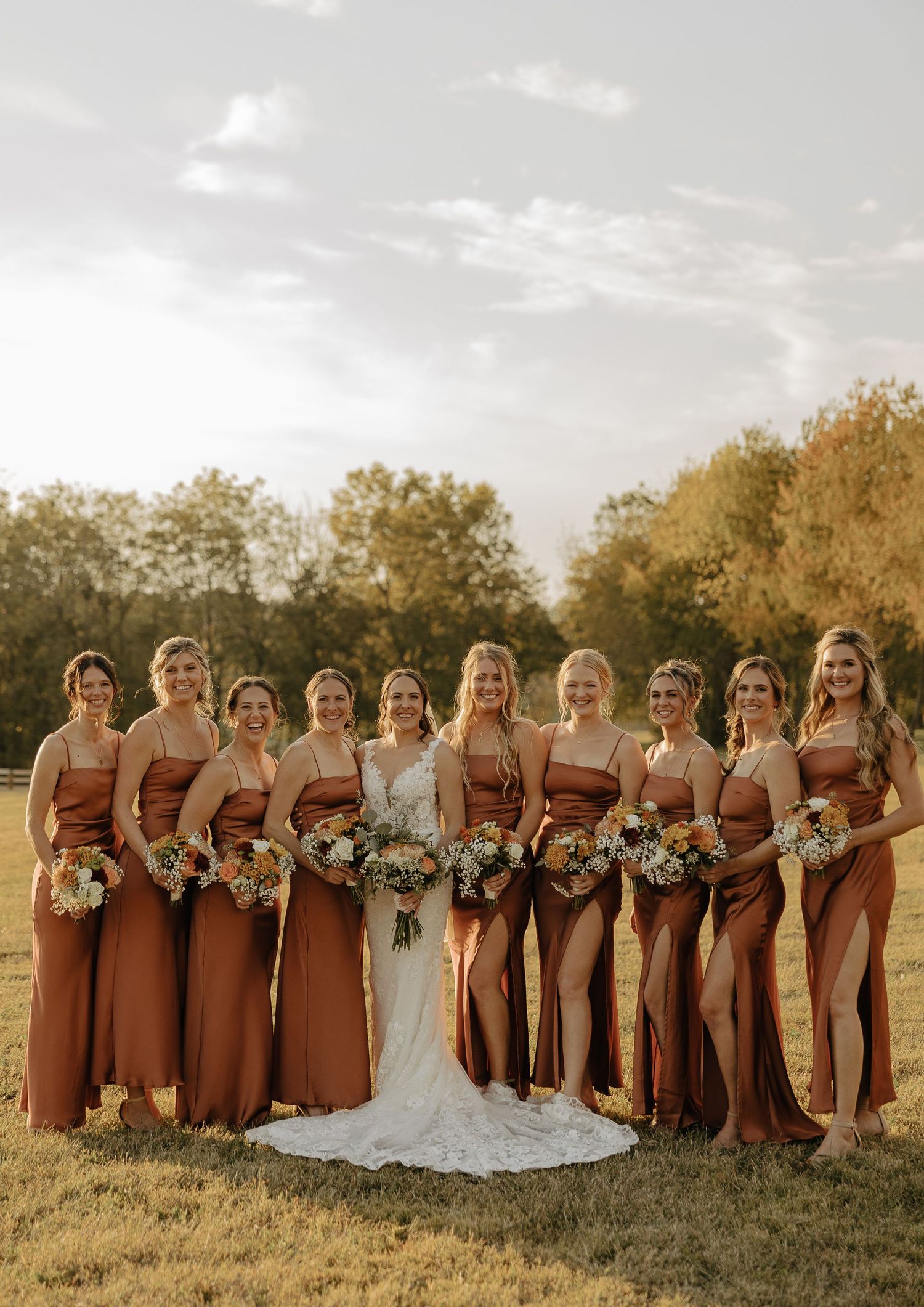Wedding party in terracotta dresses posing at sunset with autumn trees in background.