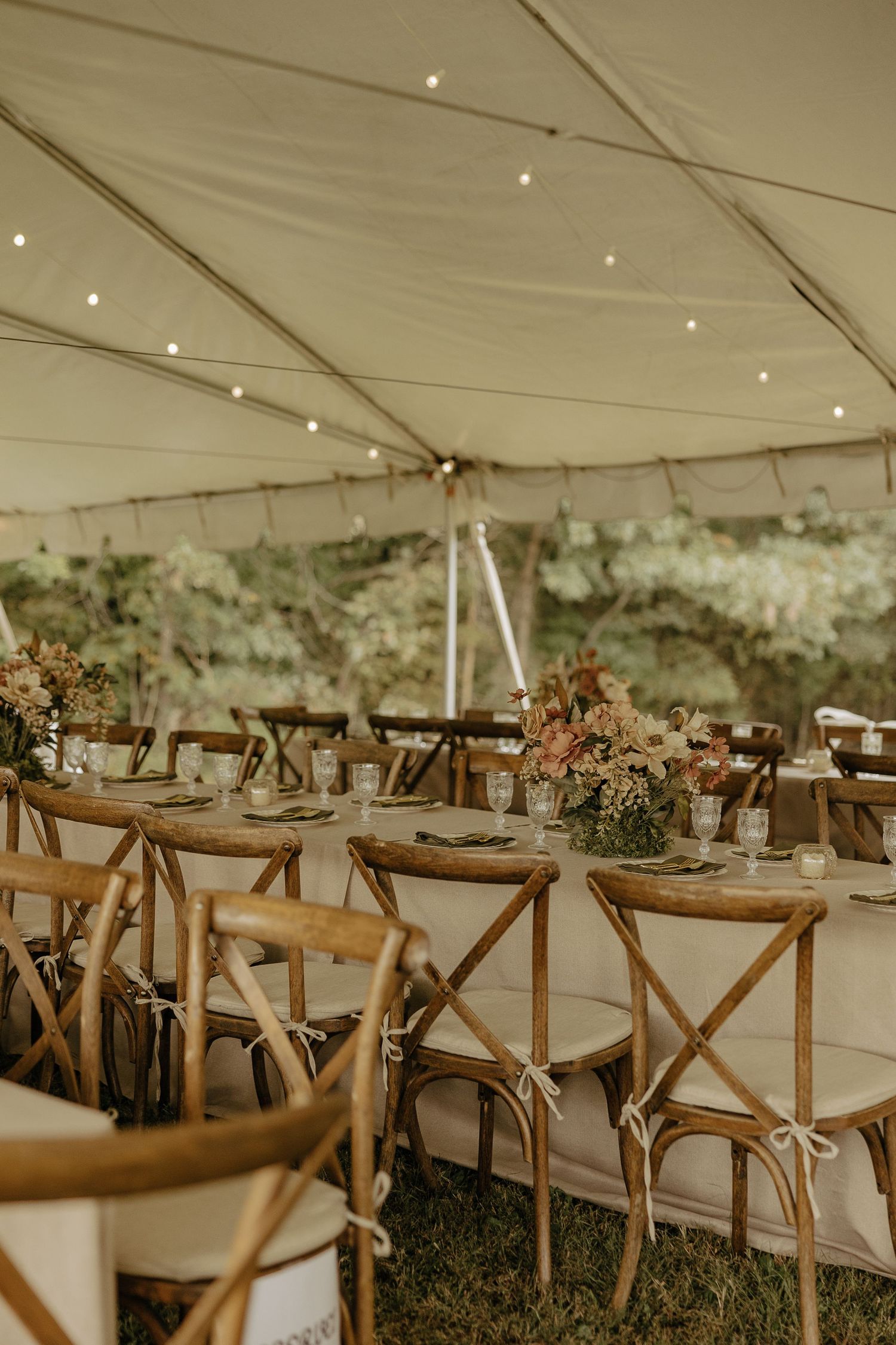 Rustic wedding reception setup under white tent with wooden cross-back chairs and floral centerpieces.