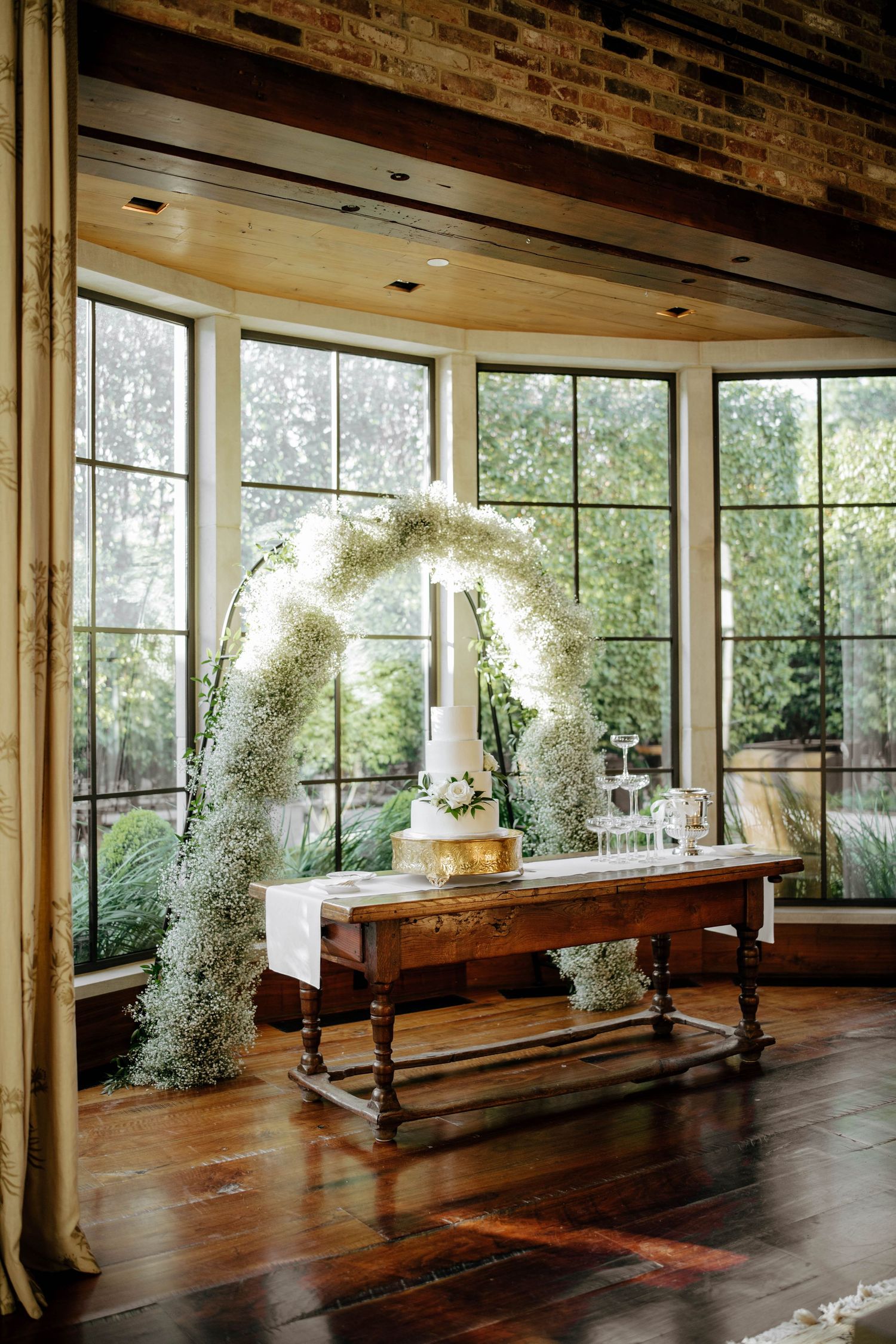Rustic wooden floor event space with floor-to-ceiling windows and elegant white floral arch decorations for a wedding ceremony.
