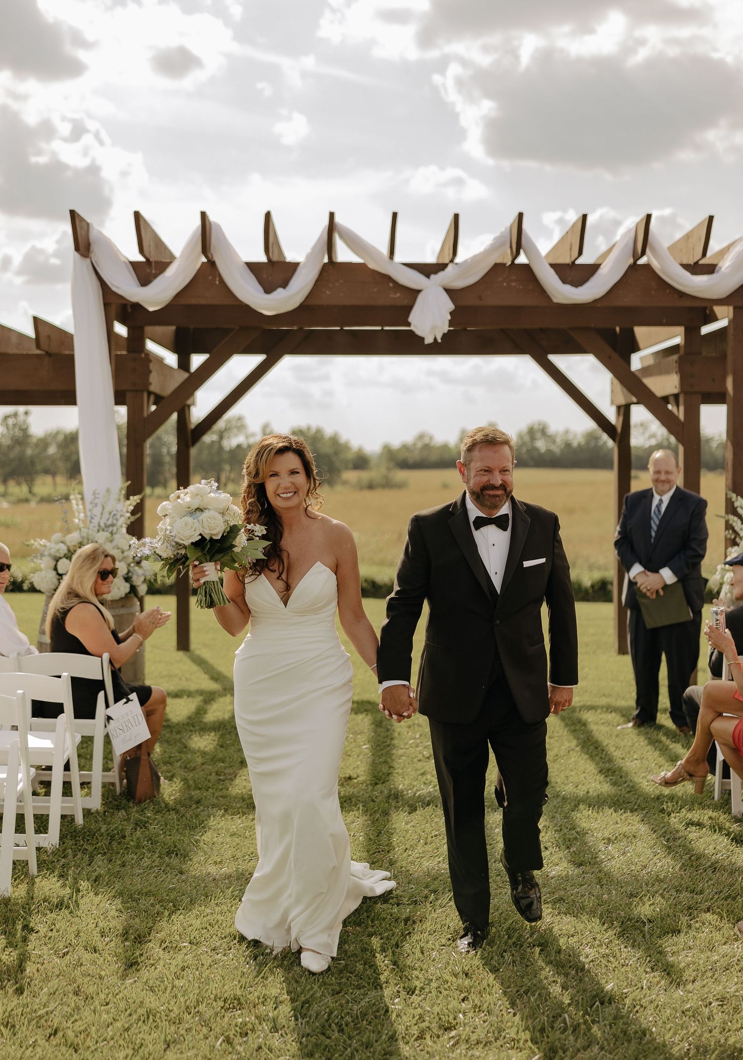 Wedding party walks joyfully down a grassy aisle under a wooden pergola decorated with white fabric on a cloudy day.