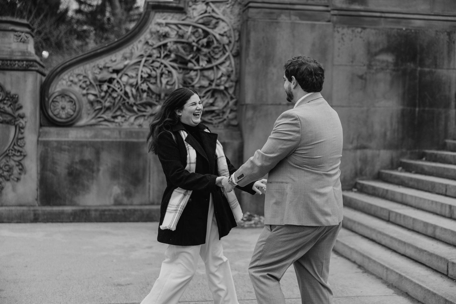 A couple shares a playful moment together near ornate stone architecture in black and white.