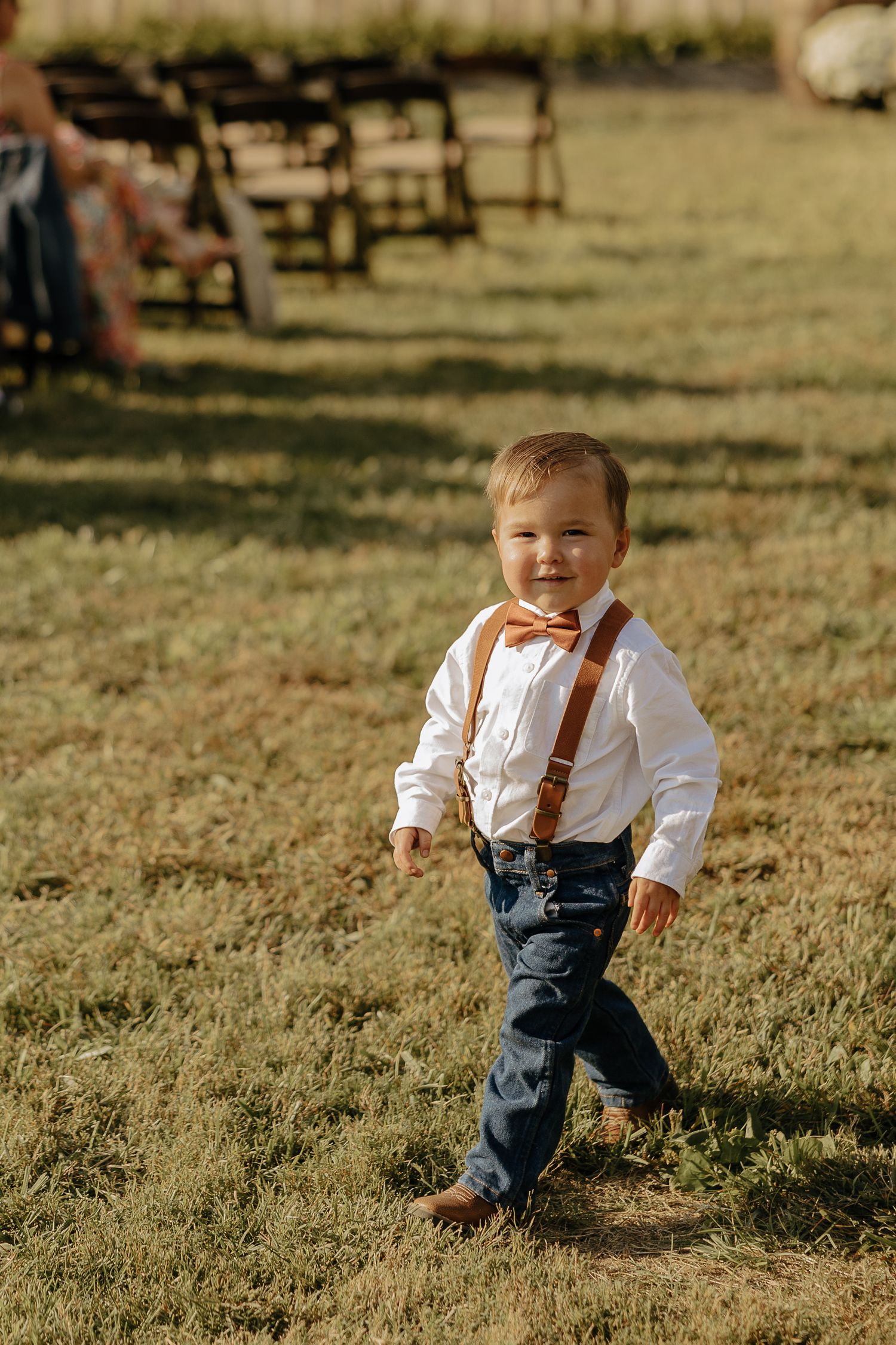 Young ring bearer in suspenders and bow tie walking on grass at outdoor wedding.