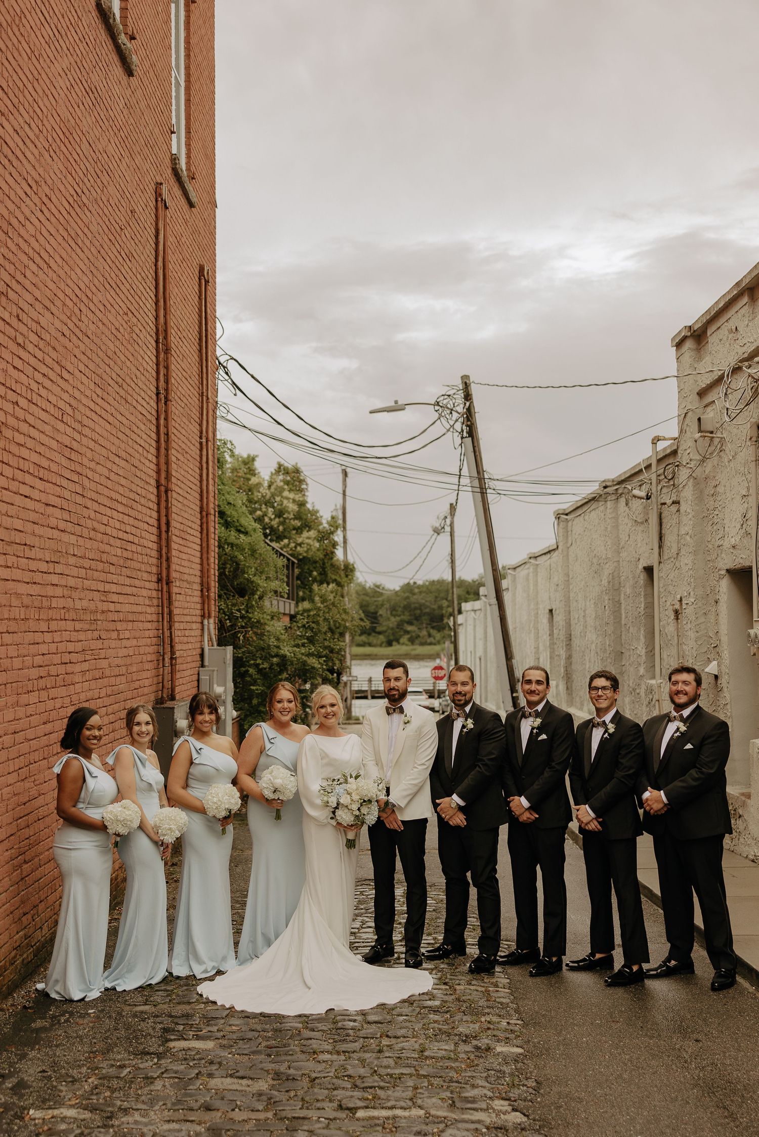 Wedding party in formal attire poses against brick wall backdrop on cobblestone street.