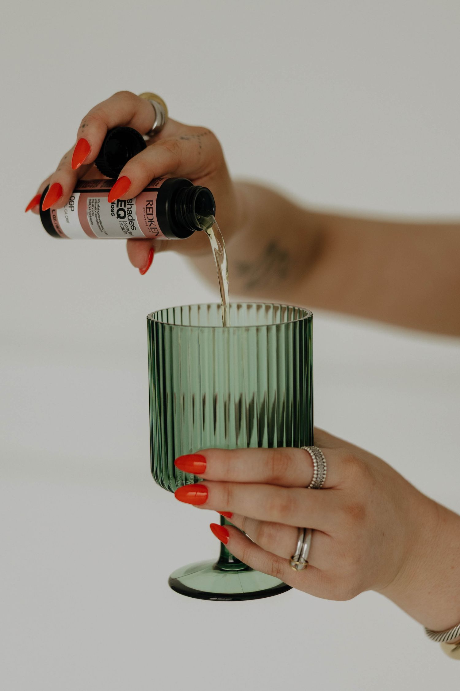 Hands with bright red nails pour liquid into a green ribbed glass goblet.