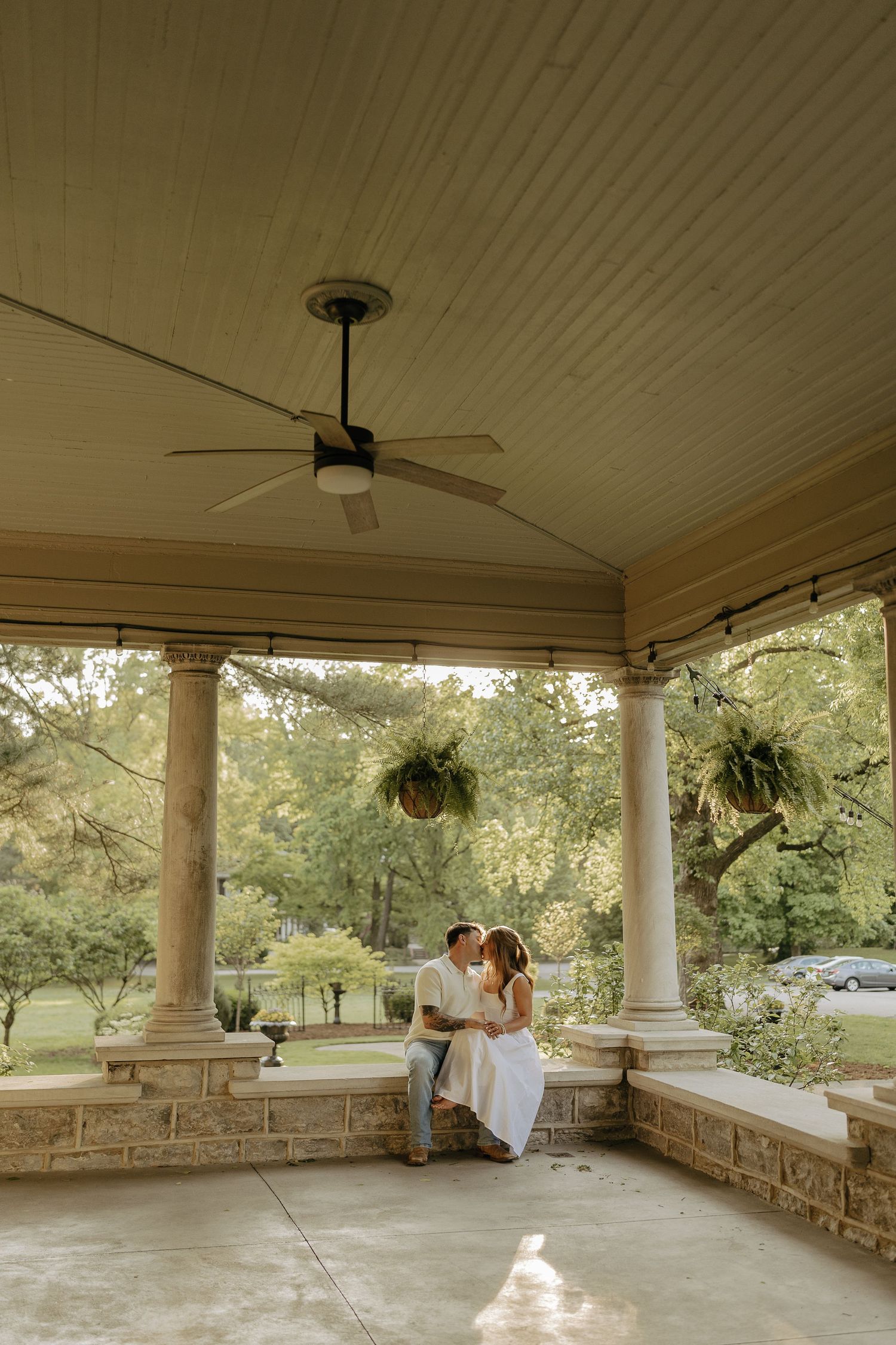 Couple sharing a romantic kiss on an elegant covered porch with columns, ceiling fan, and lush greenery in background.
