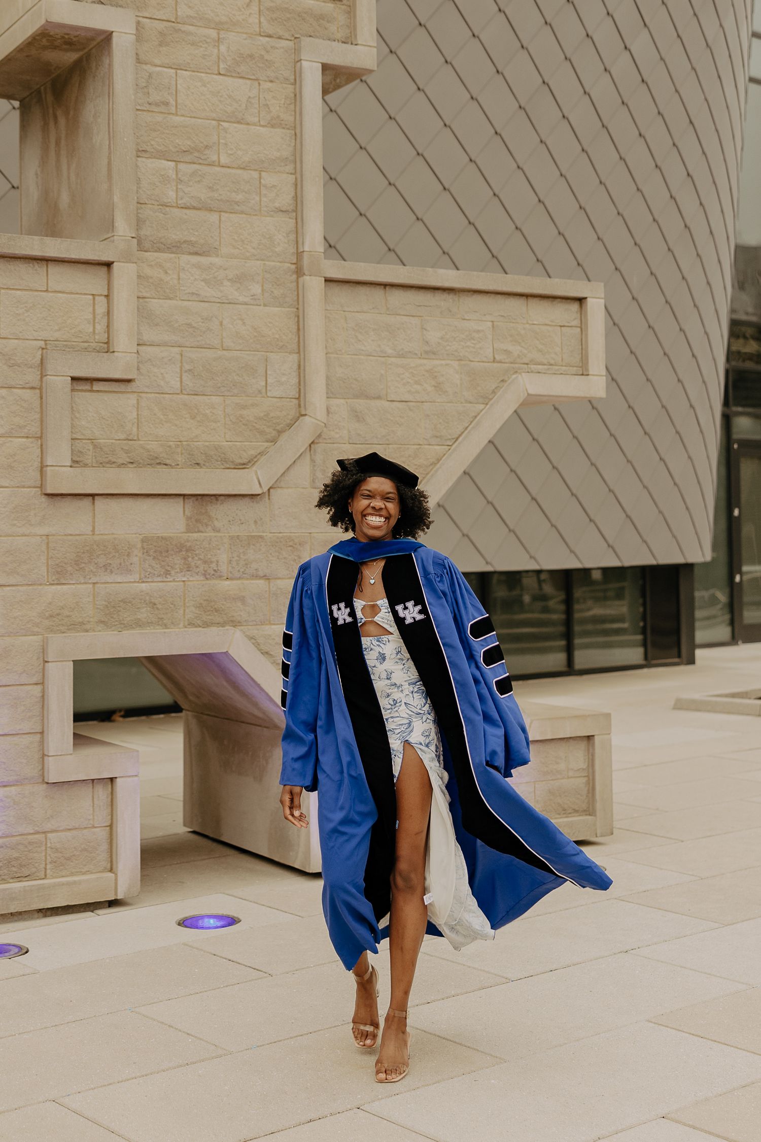 Graduate in blue doctoral regalia poses confidently against modern architectural backdrop.