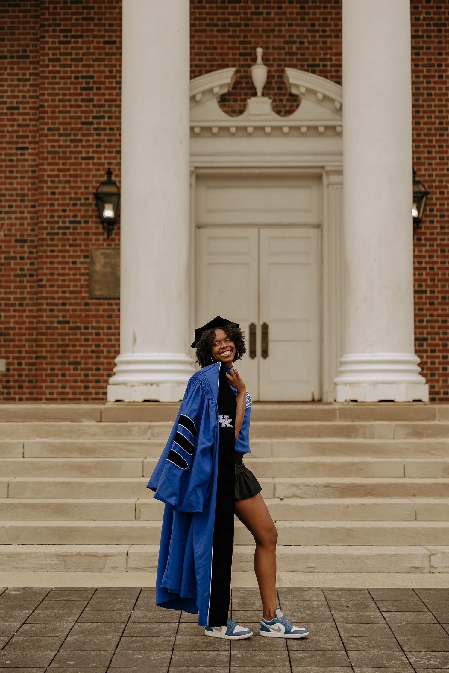 Graduate poses energetically on steps of historic brick building with white columns.
