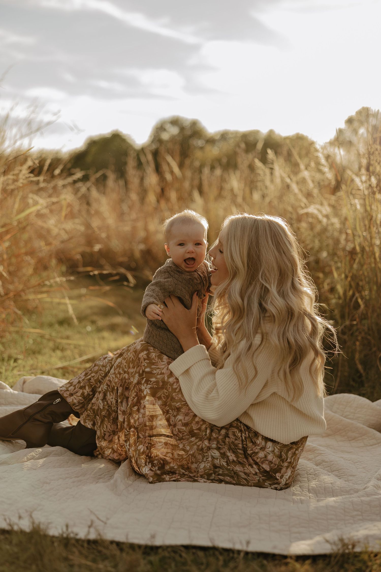 Mother and baby sharing joyful moments on white blanket during sunset photoshoot.